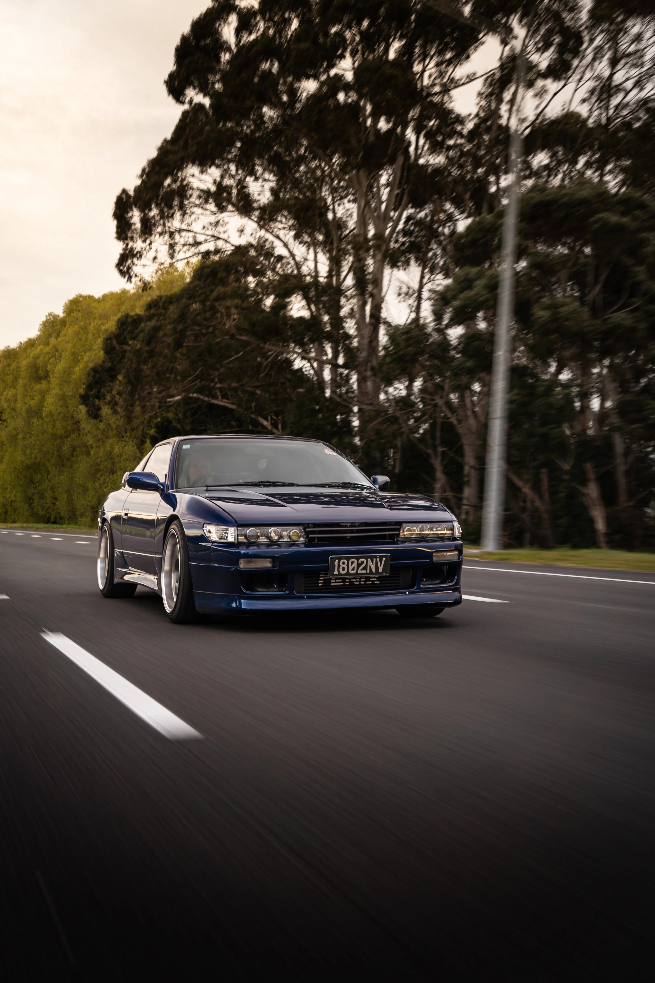 A dark blue vintage sports car driving on an open road with trees in the background.