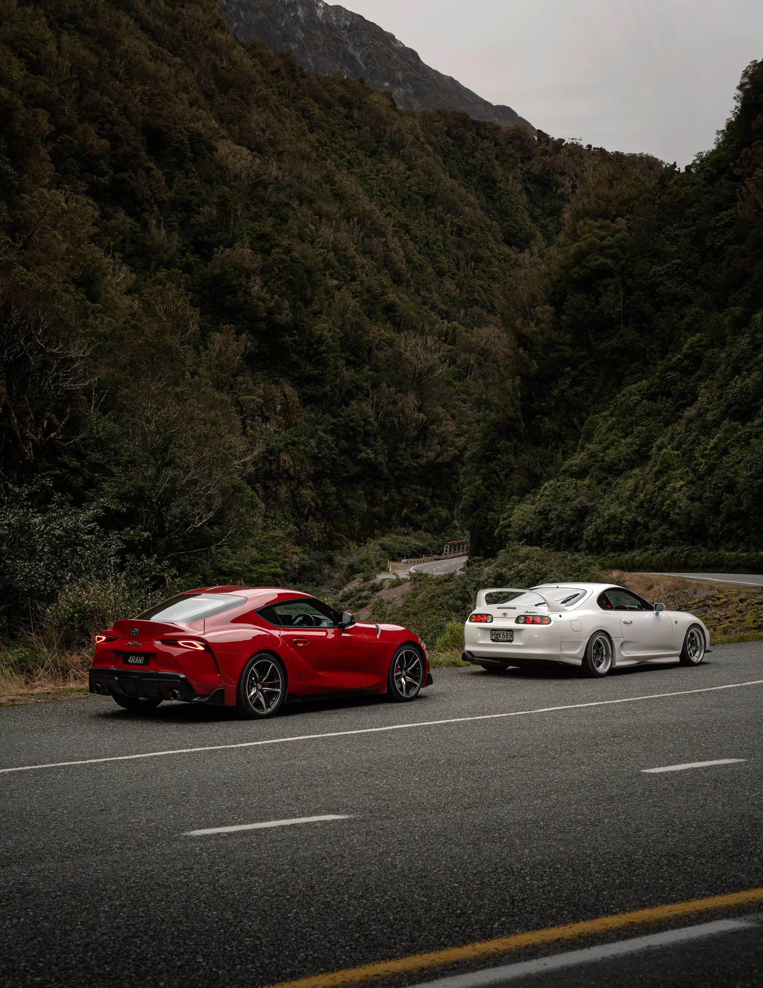 Two sports cars, one red and one white, are parked on the side of a mountain road with lush green forest and hills in the background.