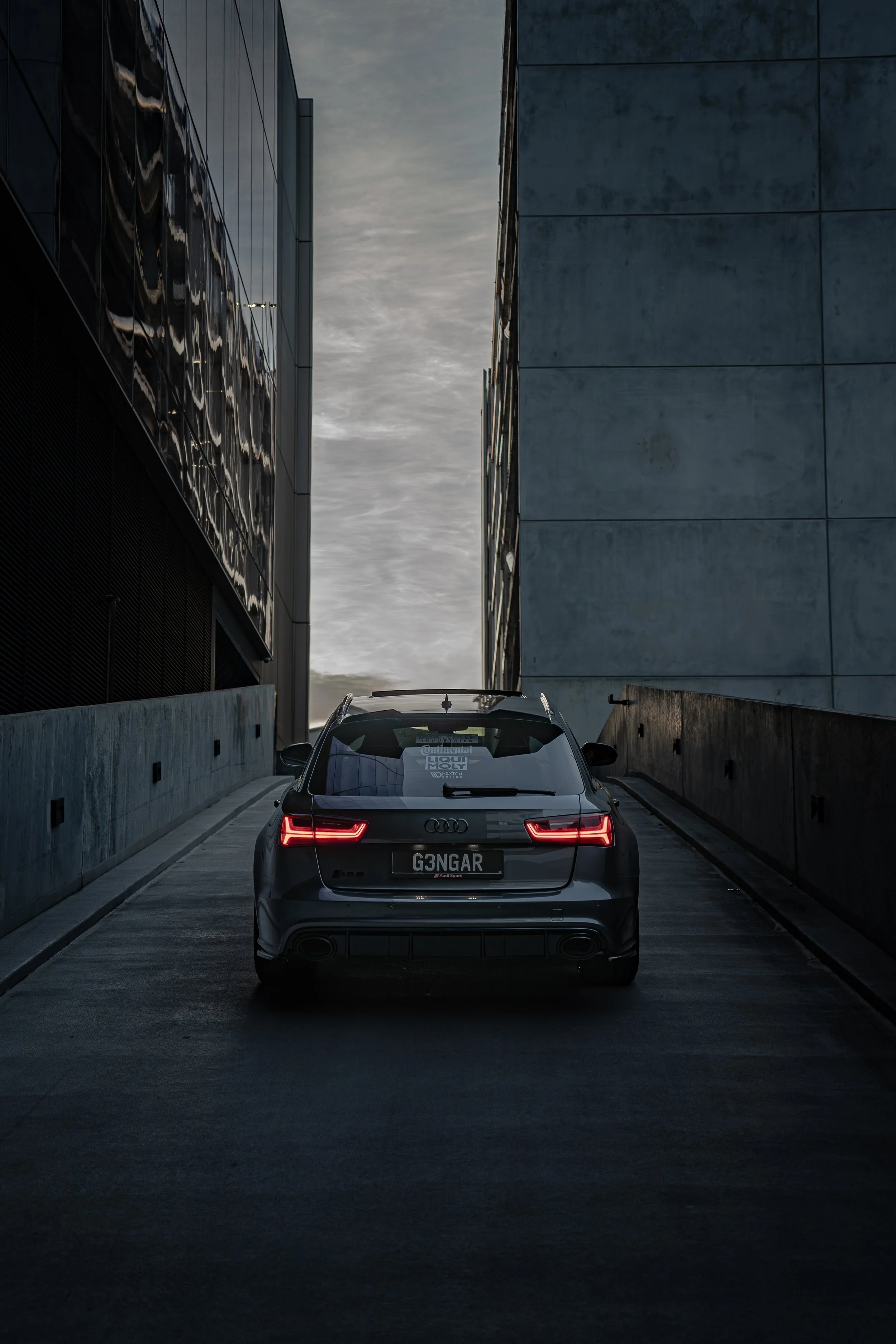 A black Audi station wagon with customized license plate 'G3NGAR' parked between two modern concrete and glass buildings at dusk.