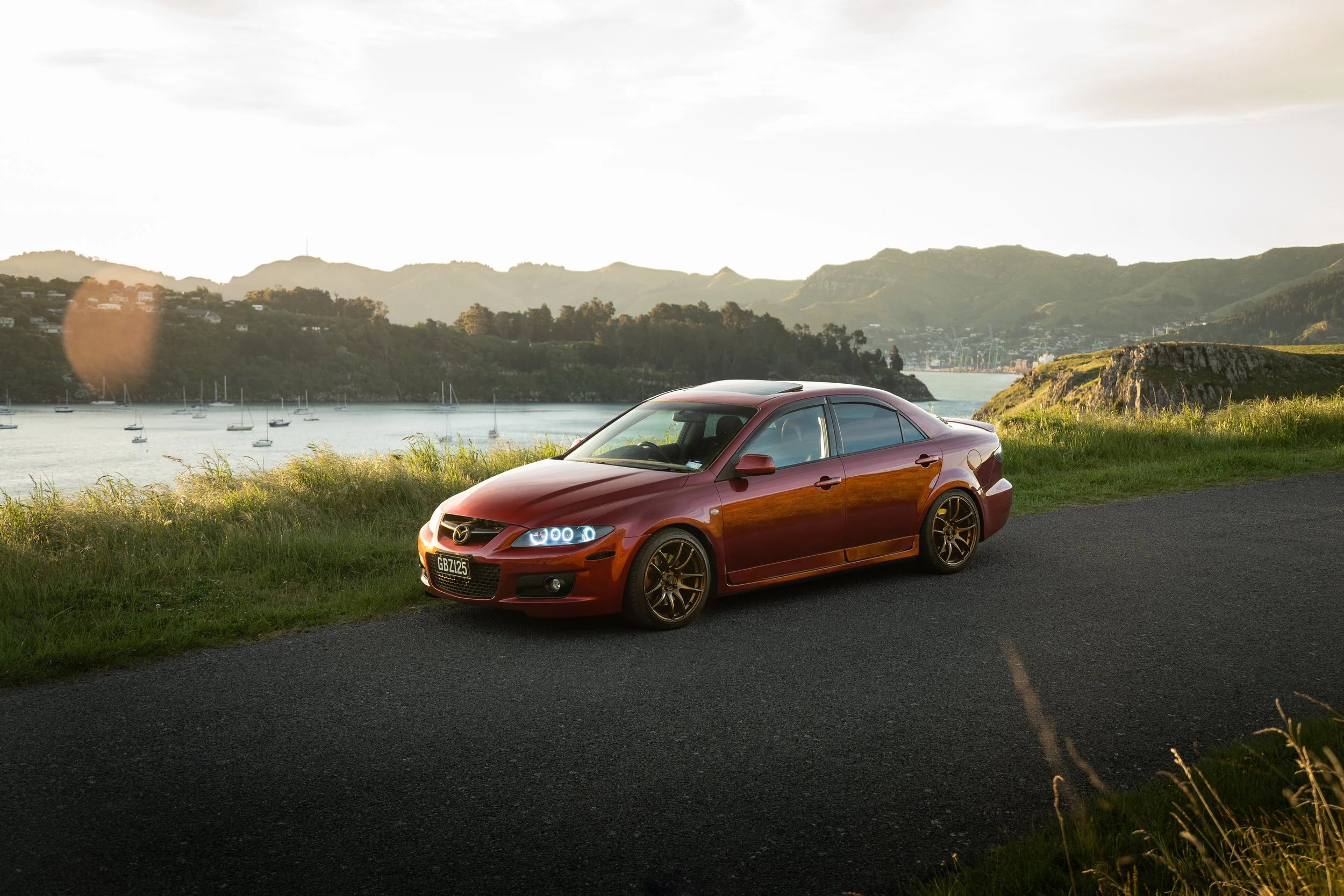 A red Mazda sedan parked beside a grassy area overlooking a body of water with sailboats, mountains, and a cloudy sky in the background.