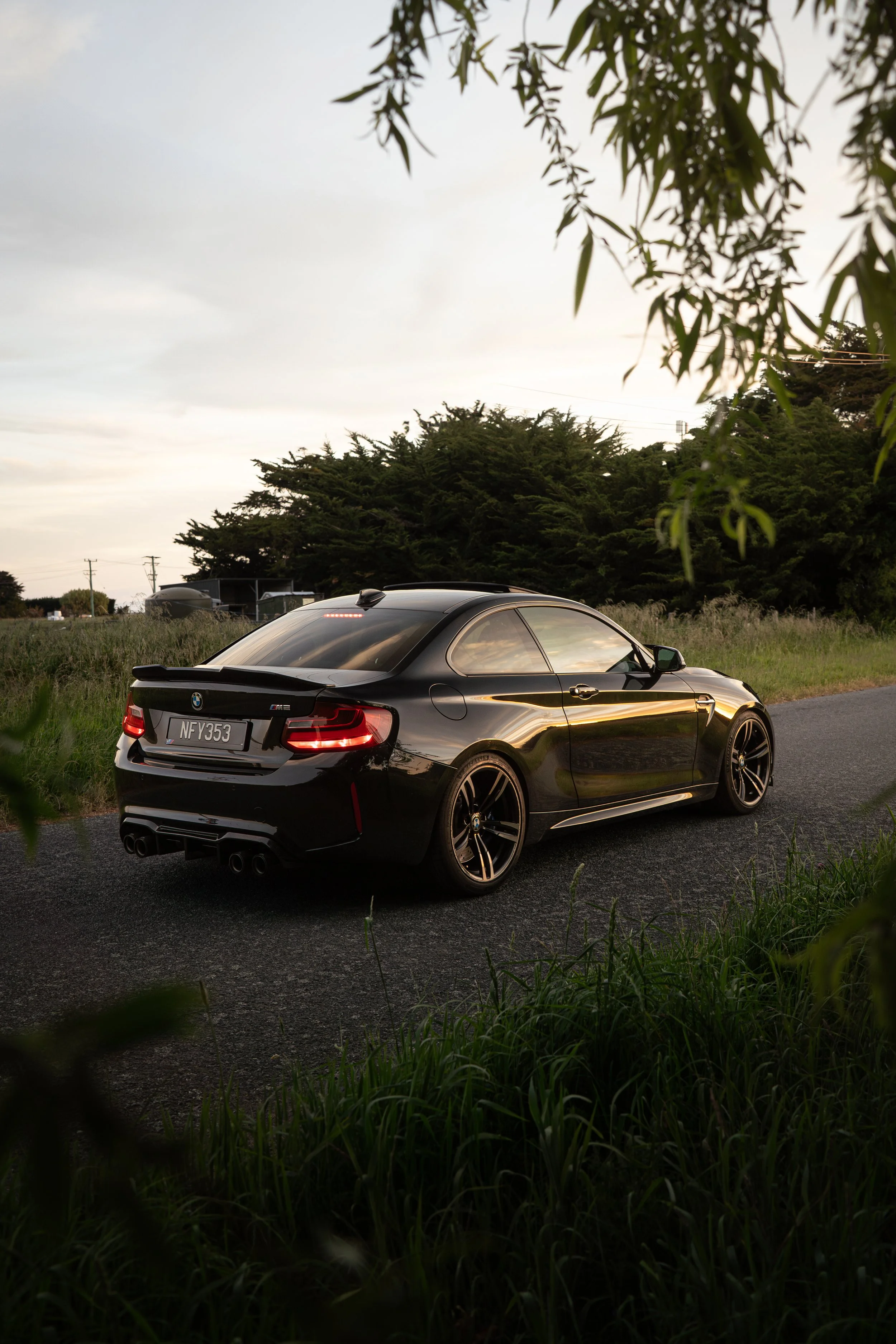 A black BMW M2 coupe parked on the side of a rural road during dusk, surrounded by grass and trees.