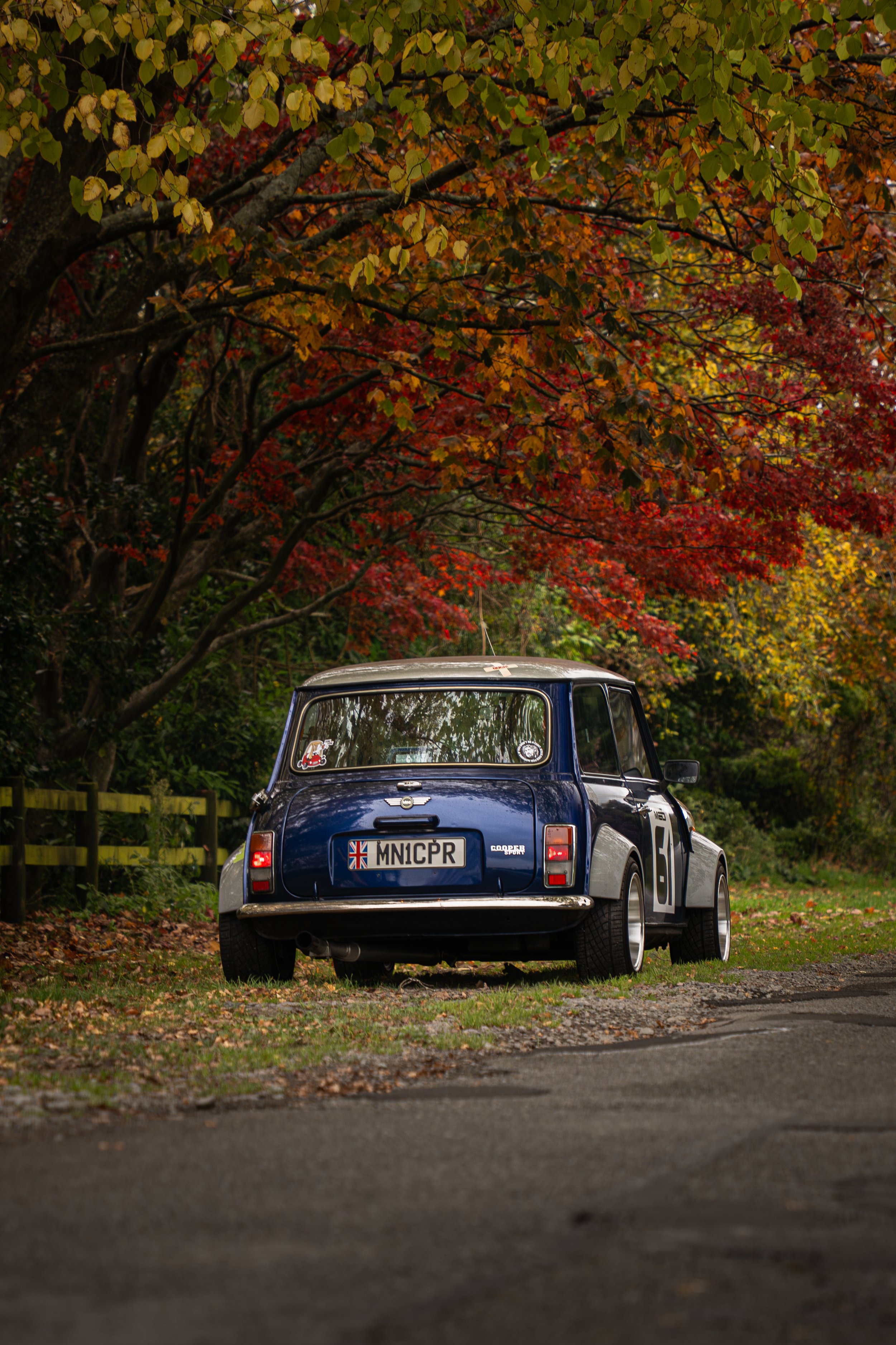 A classic blue Mini Cooper parked on the side of a road surrounded by autumn trees with red, orange, and yellow leaves.