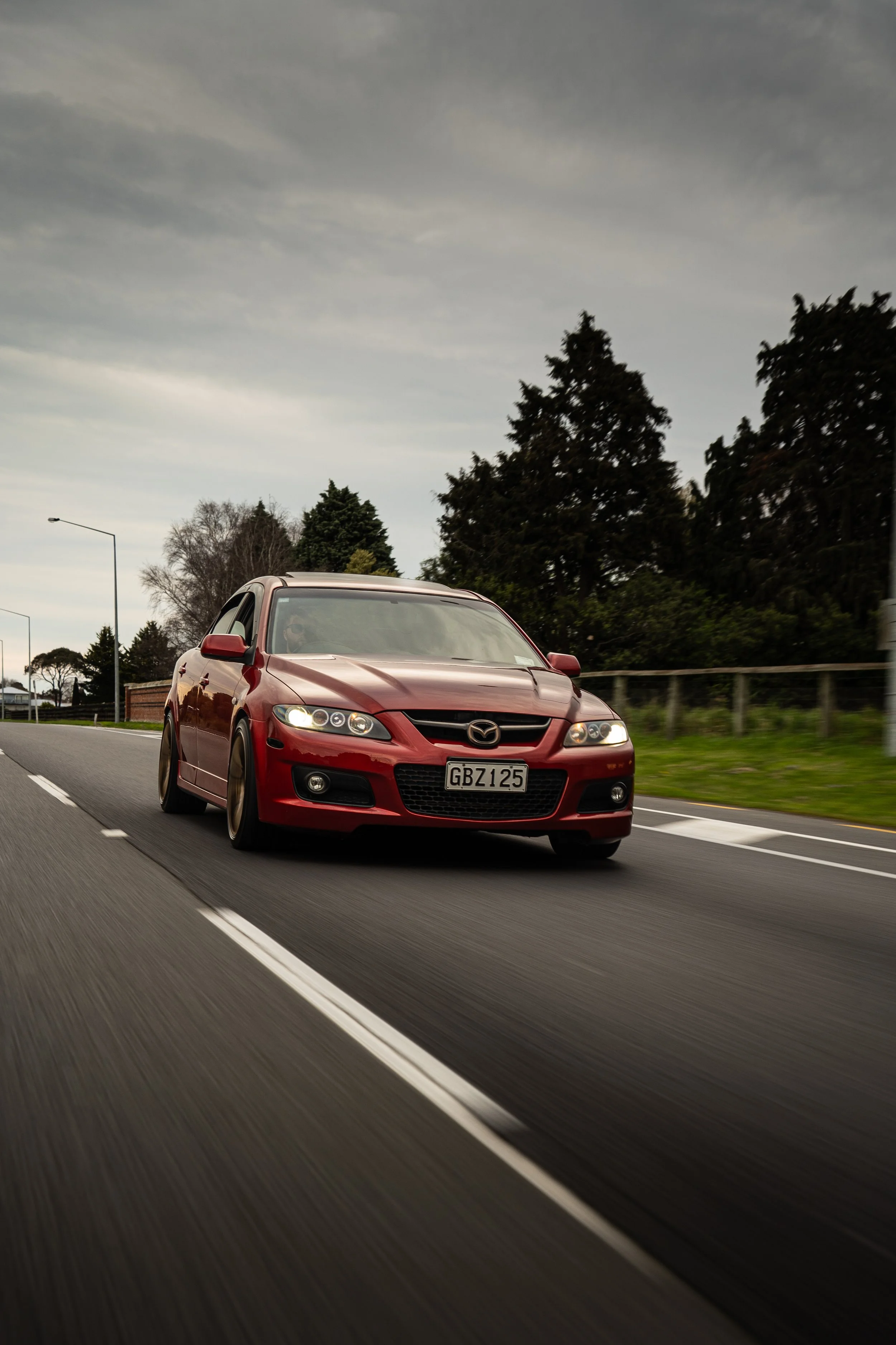 Red Mazda sports car driving on a highway with trees and cloudy sky in the background.