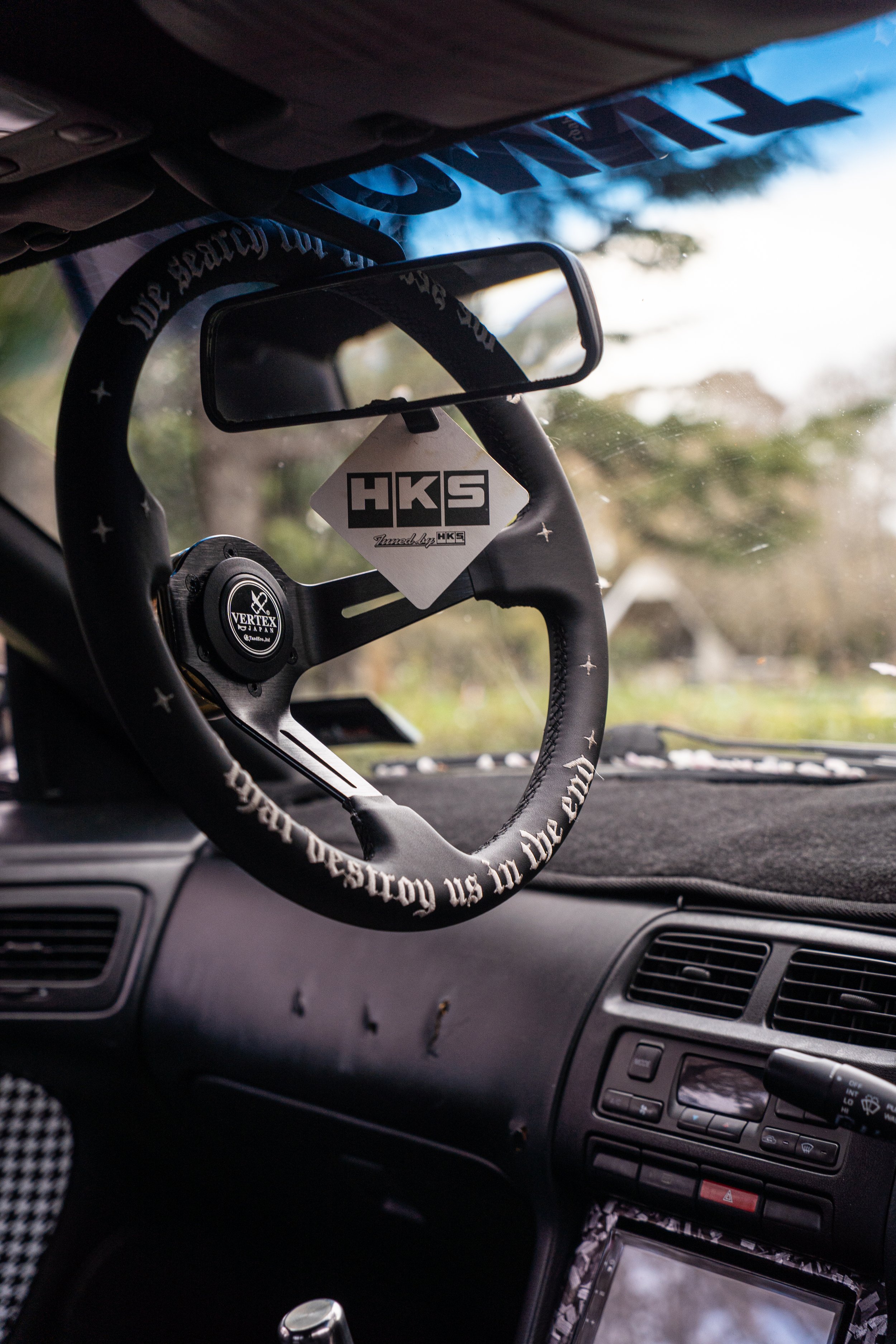 Inside a car showing a racing steering wheel with HKS tags and a Vertex Japan logo, dashboard, and windshield showing trees and sky outside.