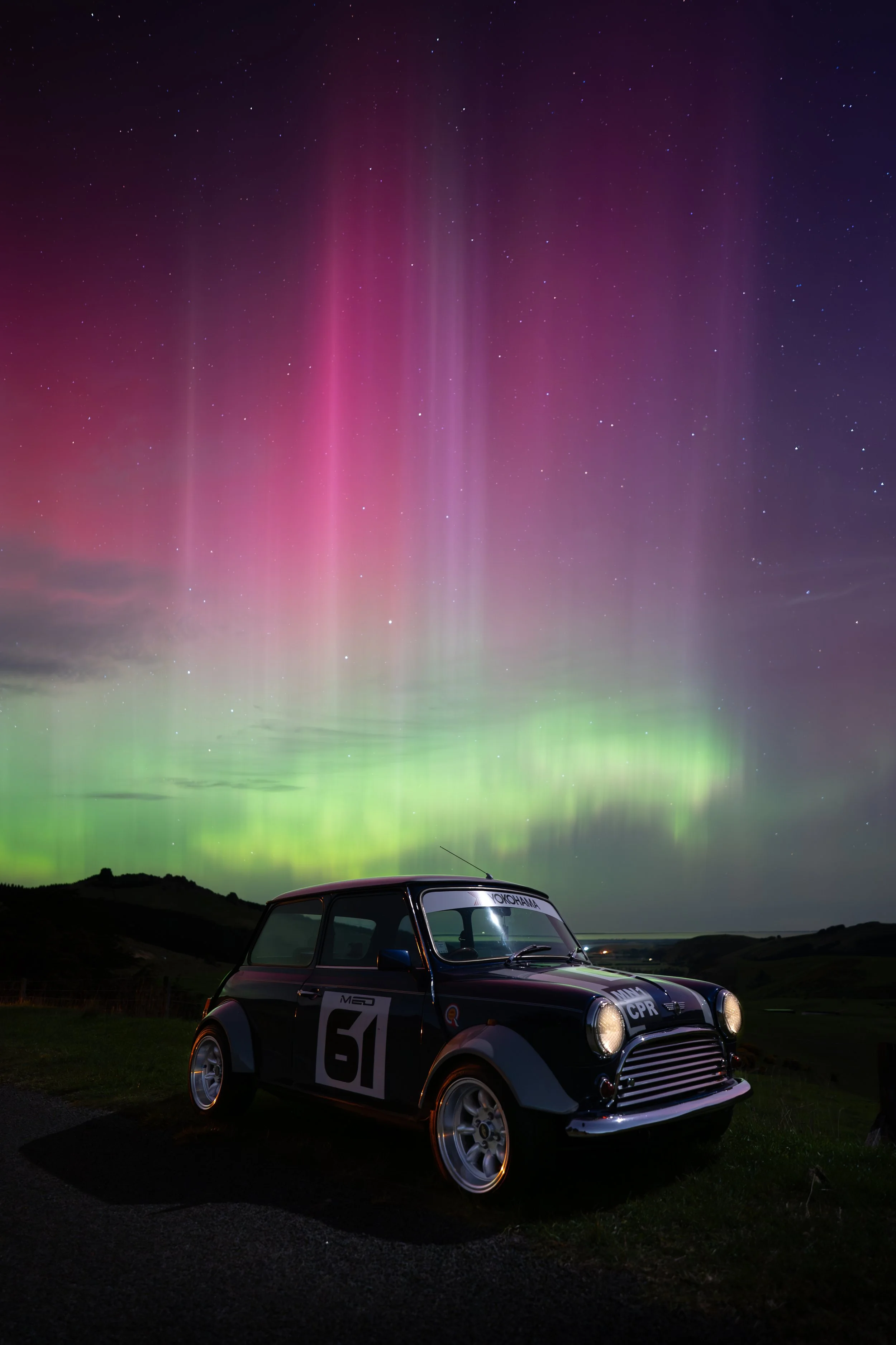 A vintage Mini Cooper car parked on a grassy area at night, with the northern lights and stars visible in the sky above.