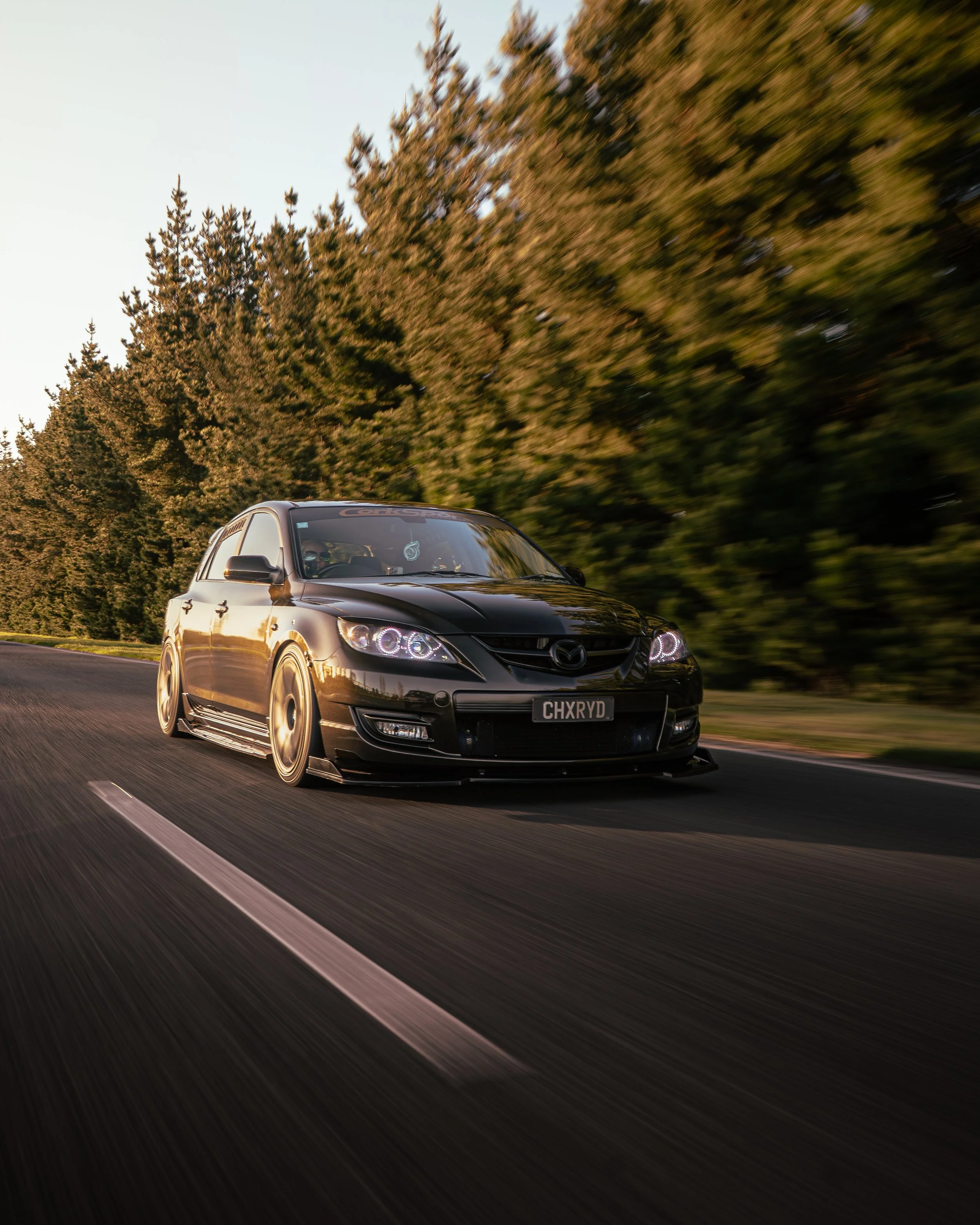 Black car racing on a road with a blurred background of trees and sky.