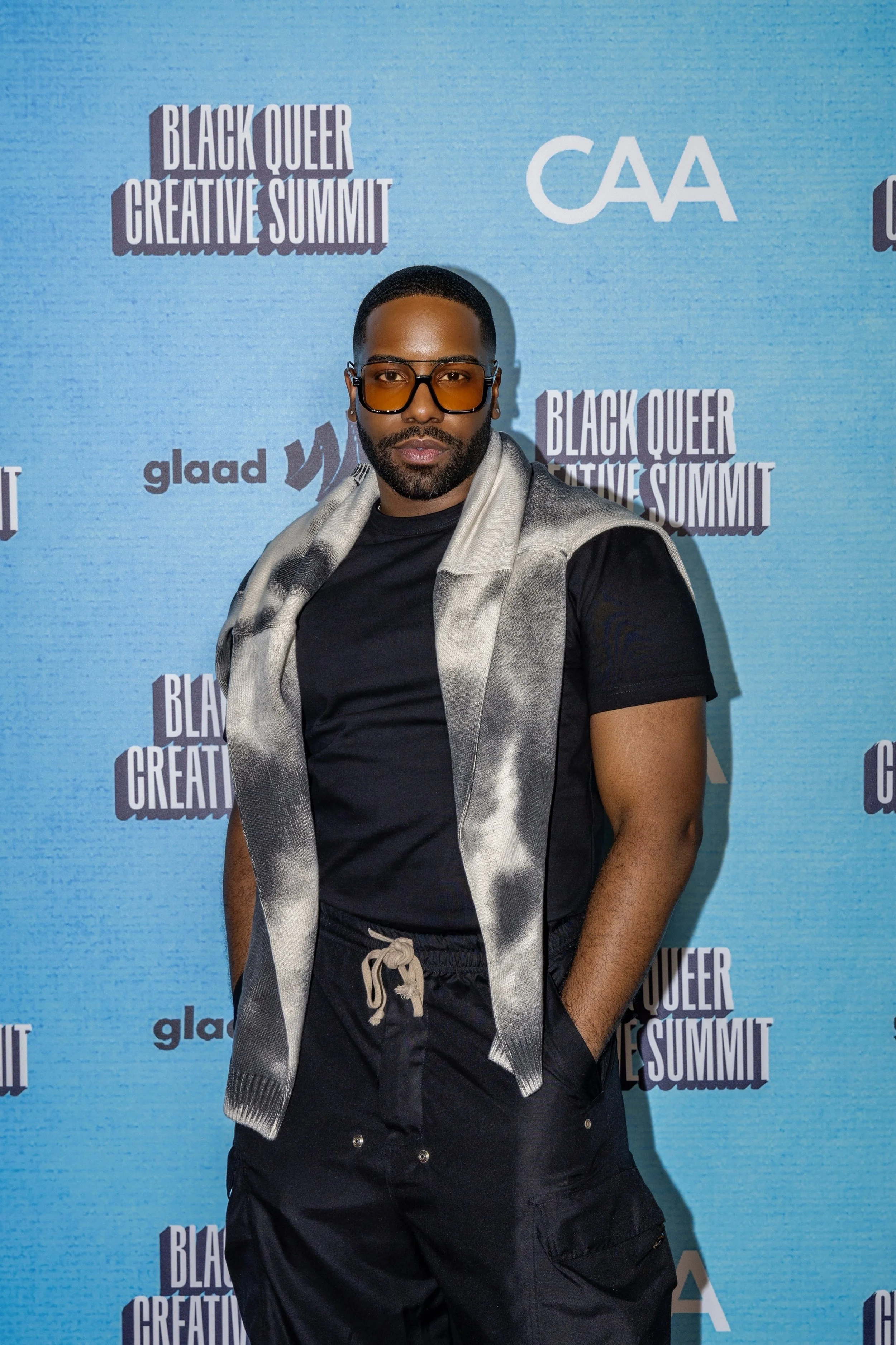 A man posing in front of a blue backdrop at the Black Queer Creative Summit, wearing sunglasses, a black t-shirt, and black pants with a gray tie-dye jacket draped over his shoulders.