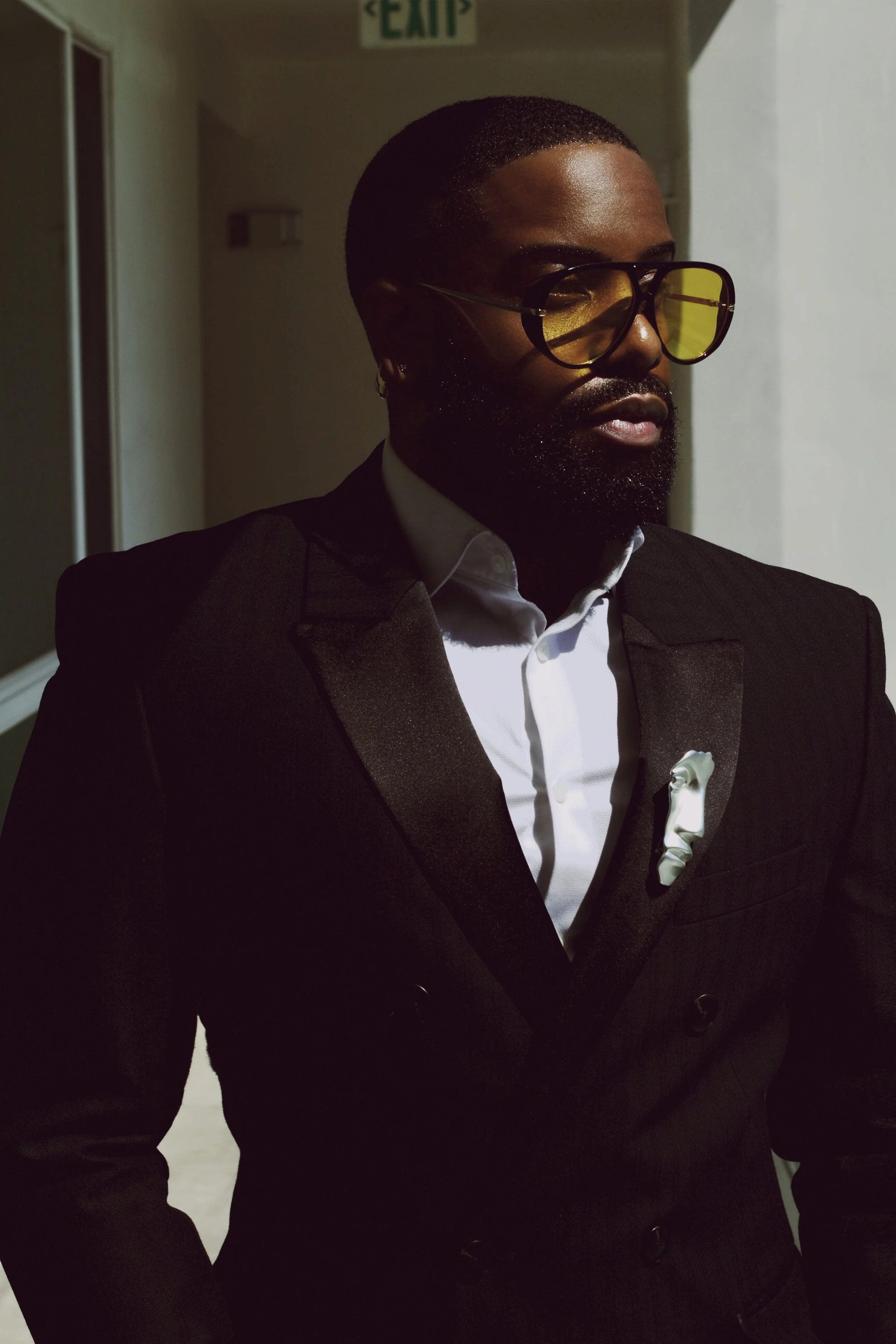 A man with a beard and glasses wearing a black tuxedo, white shirt, and a white pocket square, standing in a dimly lit hallway.