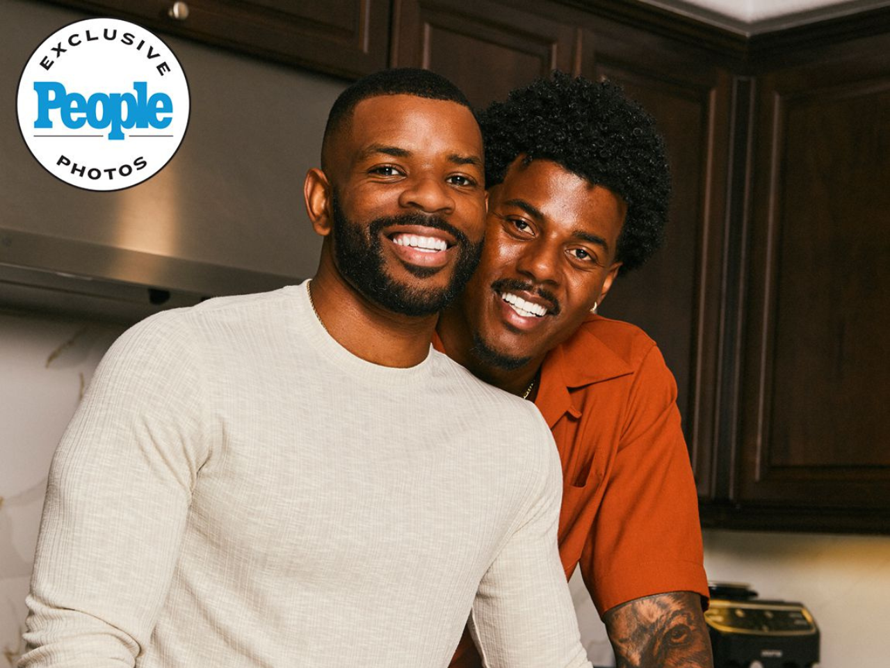 Two men smiling and leaning close together in a kitchen, one wearing a white shirt and the other an orange shirt.