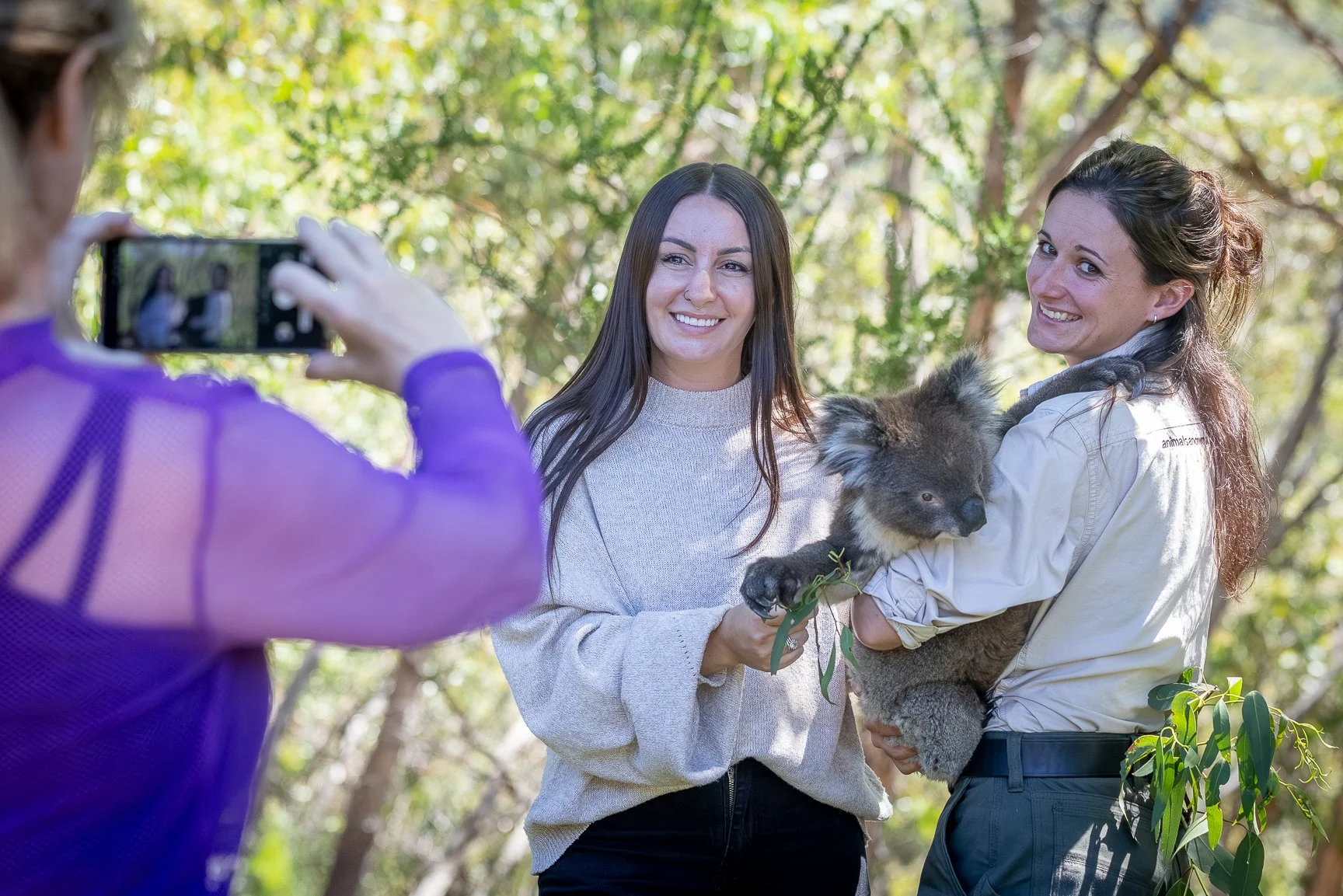 Two women are smiling and posing for a photo in a forested area, one holding a koala, while another person in a purple jacket takes the picture with a smartphone.