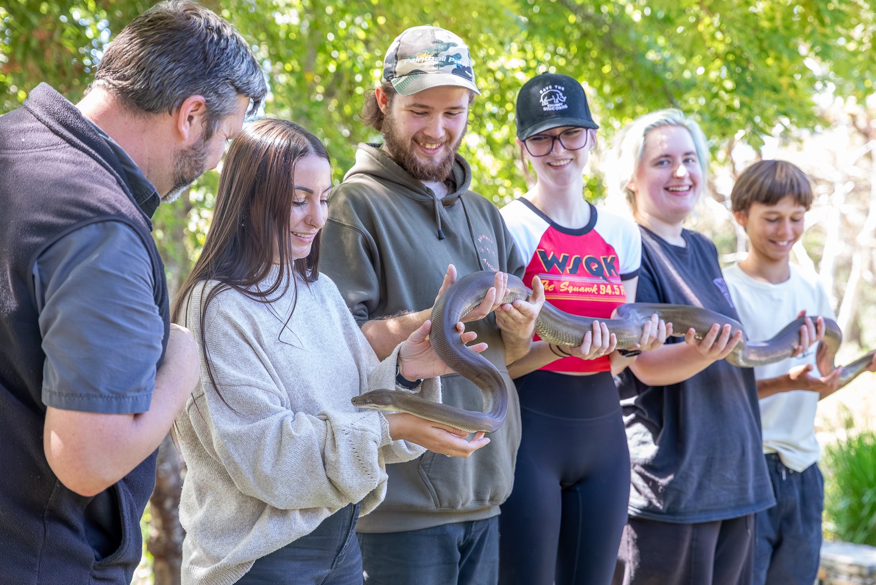Group of six people outdoors holding a carpet python, smiling, with green trees in the background.