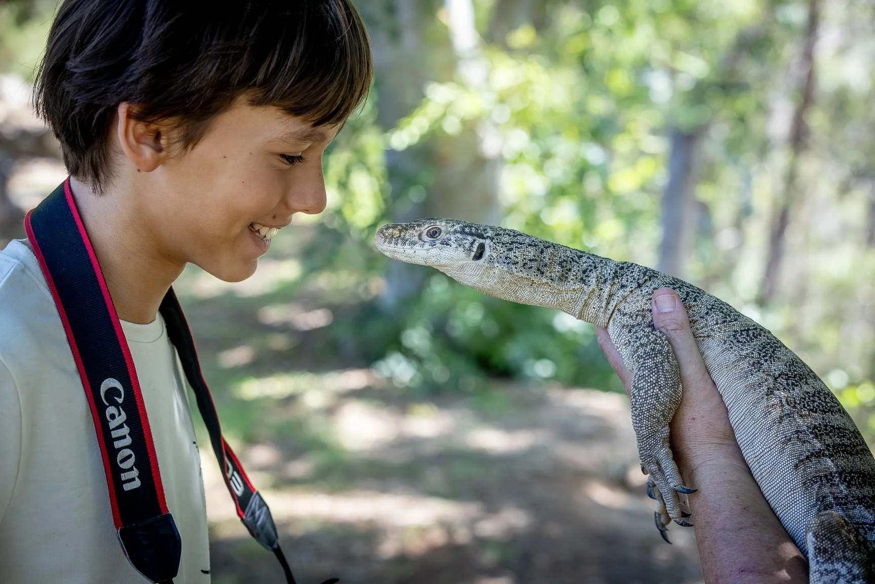 A young boy smiling while close to a goanna in a forested area.