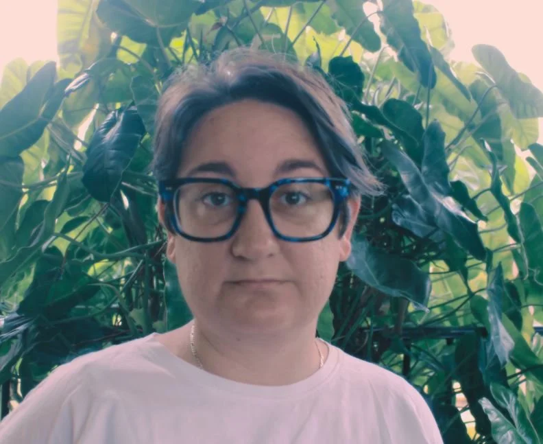 Cassandra Ramsay, a qyoung woman with short dark hair and thick blue glasses stands in the growth of a tropical rainforest.
