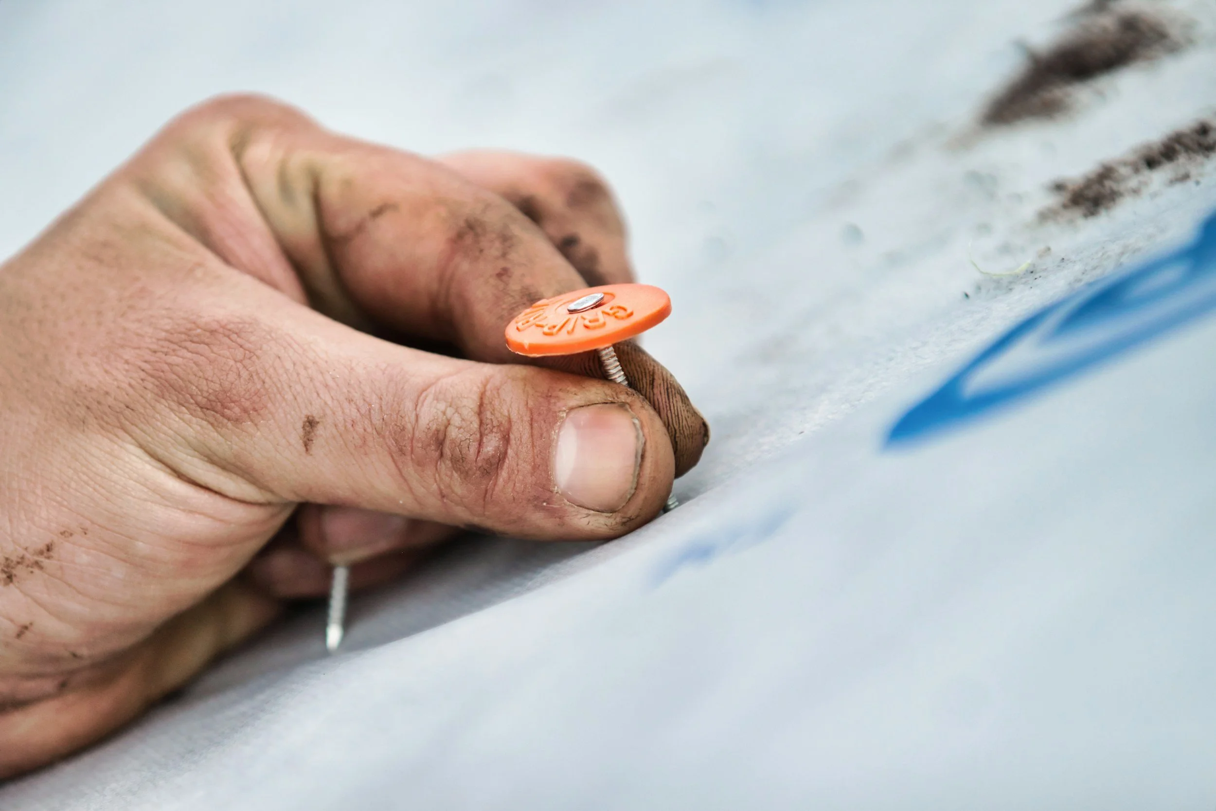 Close-up of a hand with dirt, holding a piece of climbing gear with an orange circular label, attaching it to a surface outdoors.