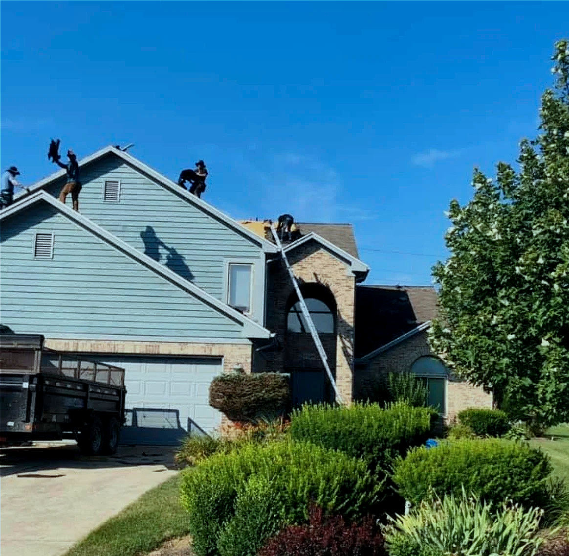 People working on the roof of a house, using ladders to access the roof, with trees, bushes, and a parked truck visible in the front yard, under a clear blue sky.