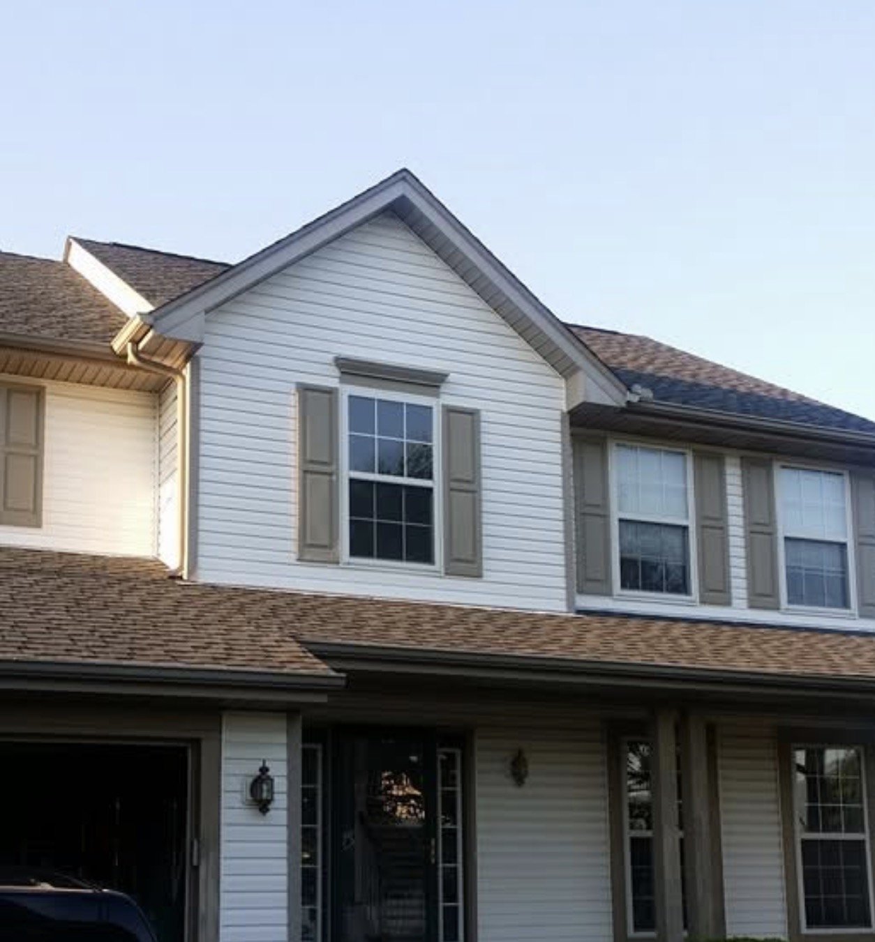 A two-story house with white siding, beige shutters, and a brown shingled roof, featuring multiple windows and a front porch area.