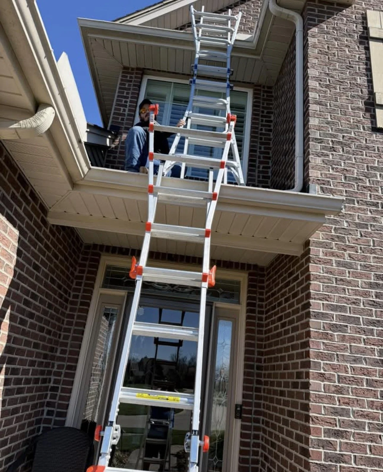 A man on a balcony with a tall extension ladder leaning against the house, working on the roof or gutter, in a residential brick home with a blue sky in the background.