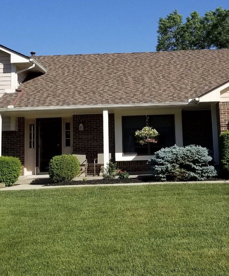 Front of a house with a brick exterior, a brown shingled roof, a covered porch with two chairs, and neatly trimmed bushes and plants in the yard.