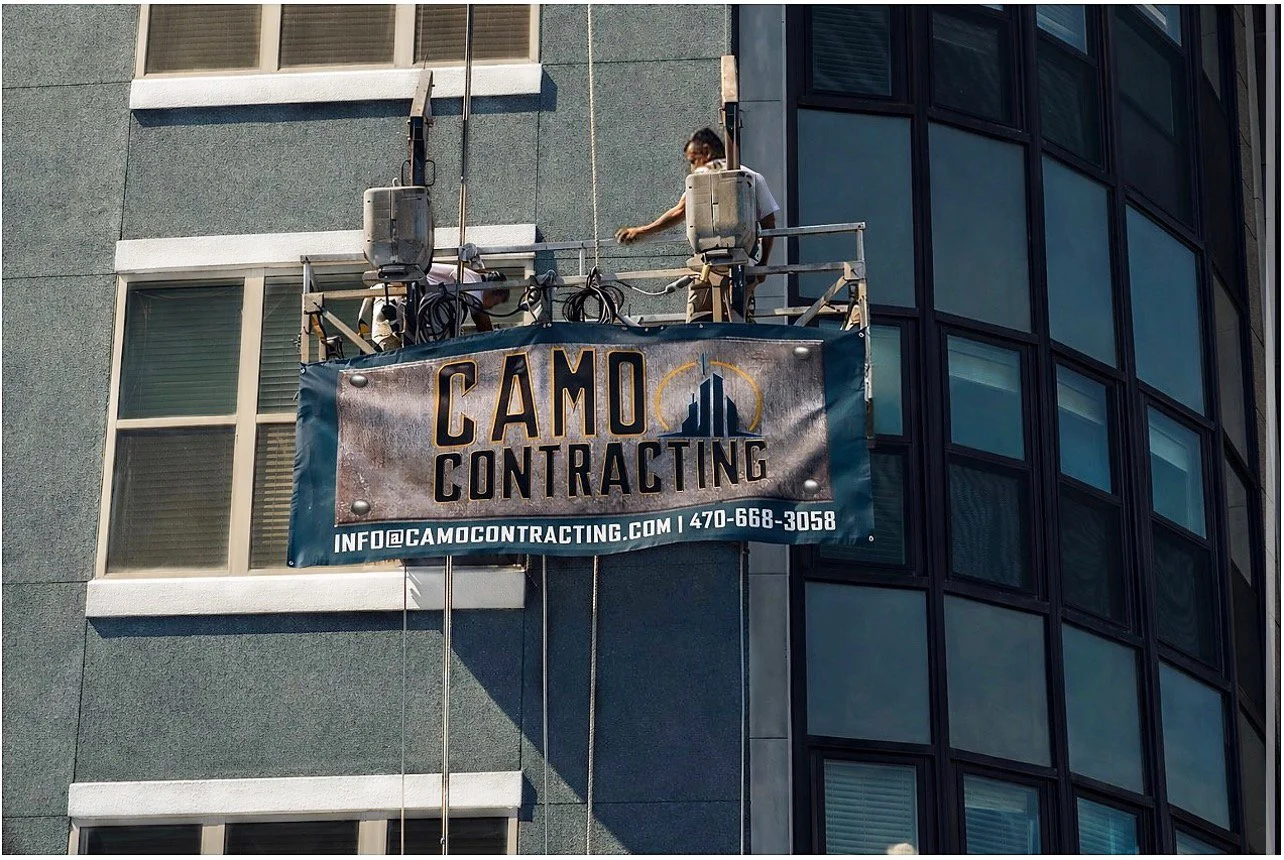 A window cleaner working on the exterior of a high-rise building using a suspended platform with a banner that reads 'Camo Contracting' and includes contact information.
