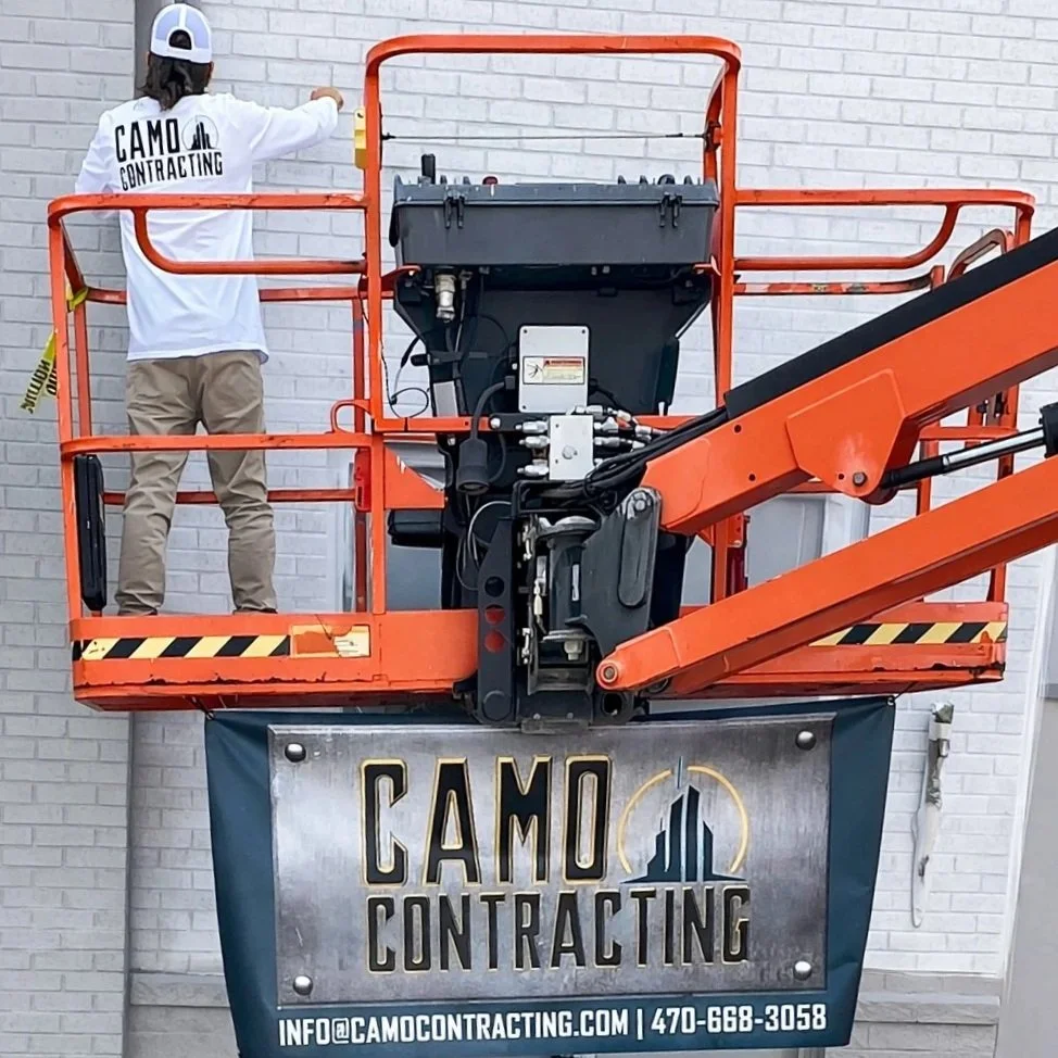 A worker on an orange lift installing or repairing a sign for CAMO Contracting on a brick wall. The sign displays the company name and contact information.