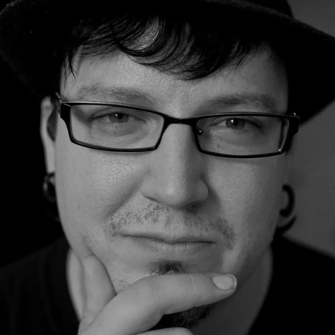 A black and white close-up of a man with glasses, dark hair, and a hand on his chin, looking contemplative.