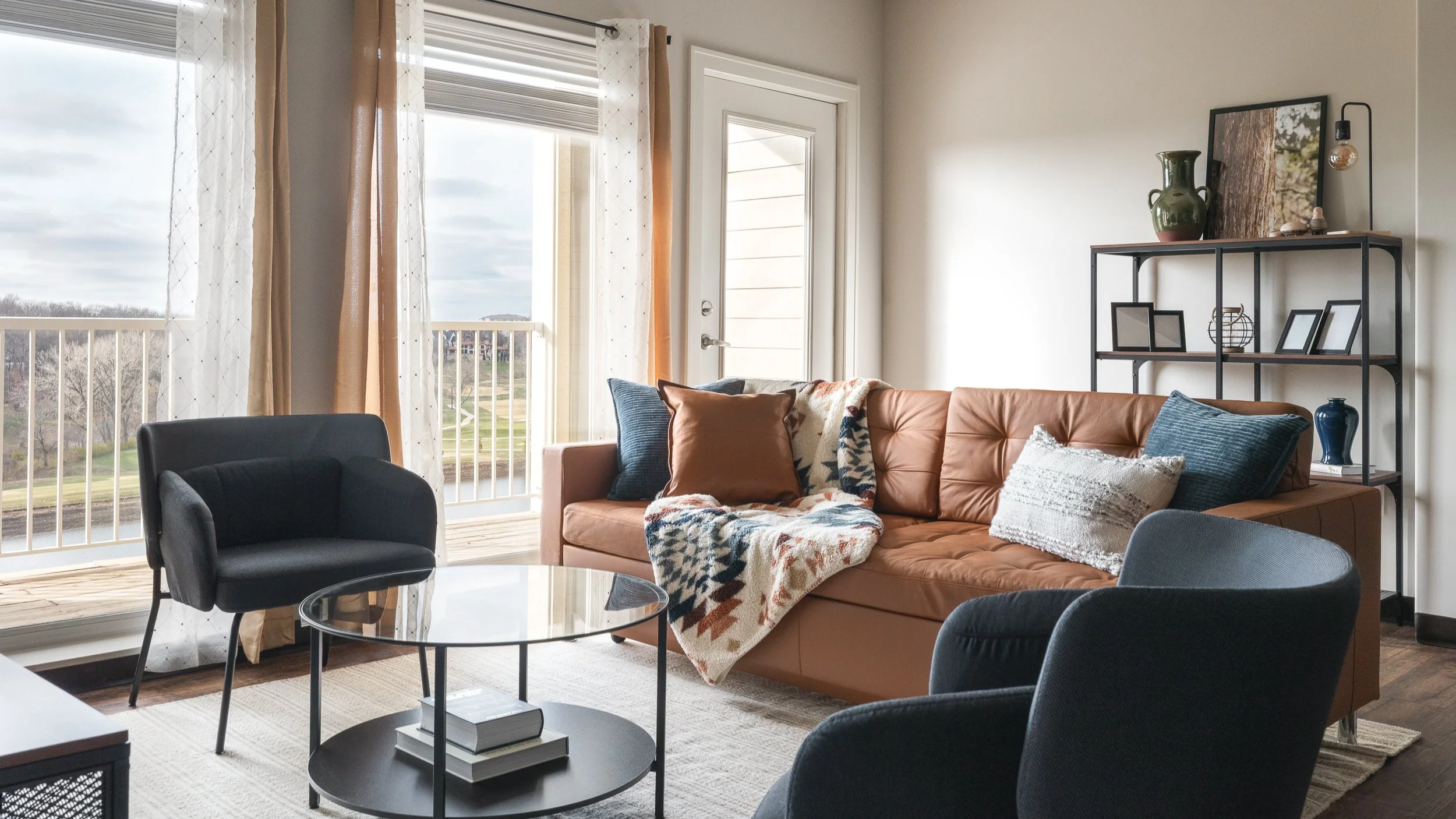Living room with a brown leather sofa, black armchair, glass coffee table, and a black metal shelf with photo frames, vases, and artwork. Large window with sheer curtains and a door leading outside, revealing an outdoor balcony with railings and a view of trees and a cloudy sky.