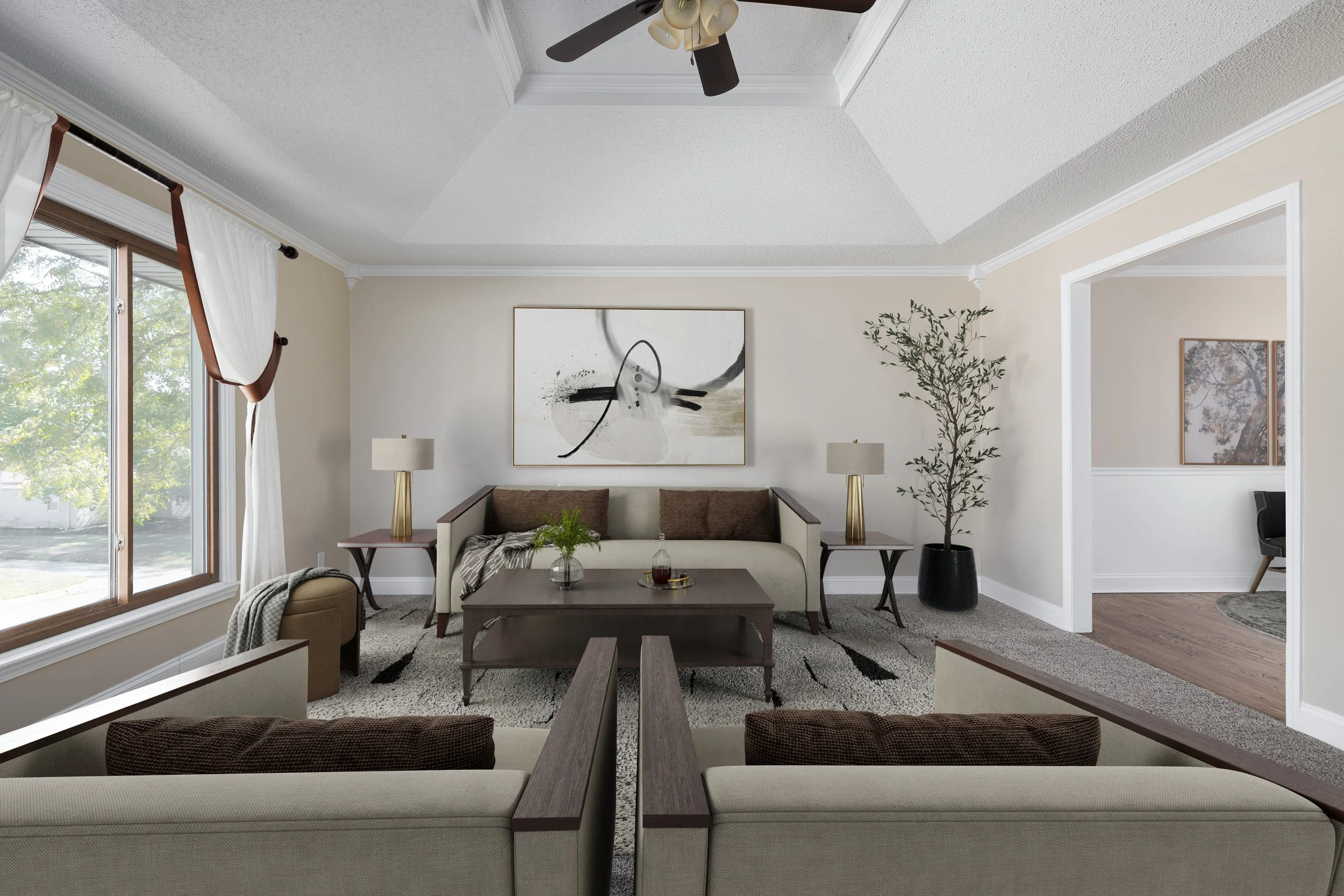 Living room with beige walls and large window with white curtains. A dark wood coffee table in front of a beige sofa, flanked by two table lamps. Abstract black and white art on the wall and a potted plant in a black planter. Ceiling fan with light fixtures.