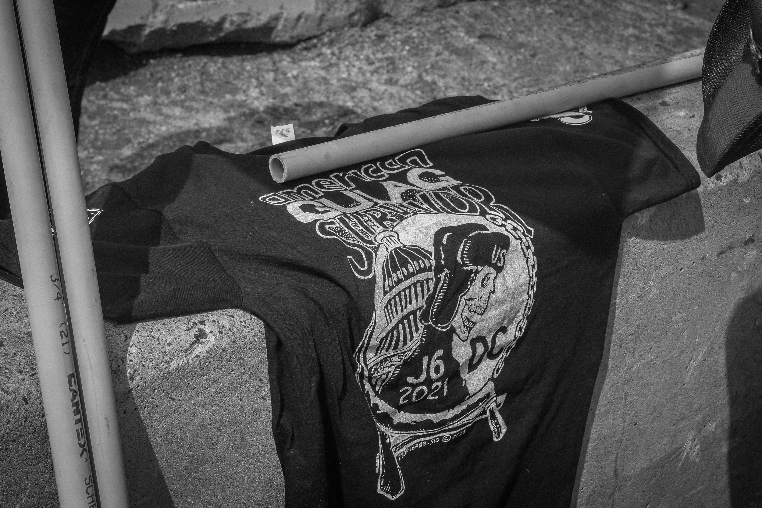 Black-and-white photo of a graphic T-shirt reading “American Ska,” with a skull and microphone design, beside PVC pipes on a concrete surface.
