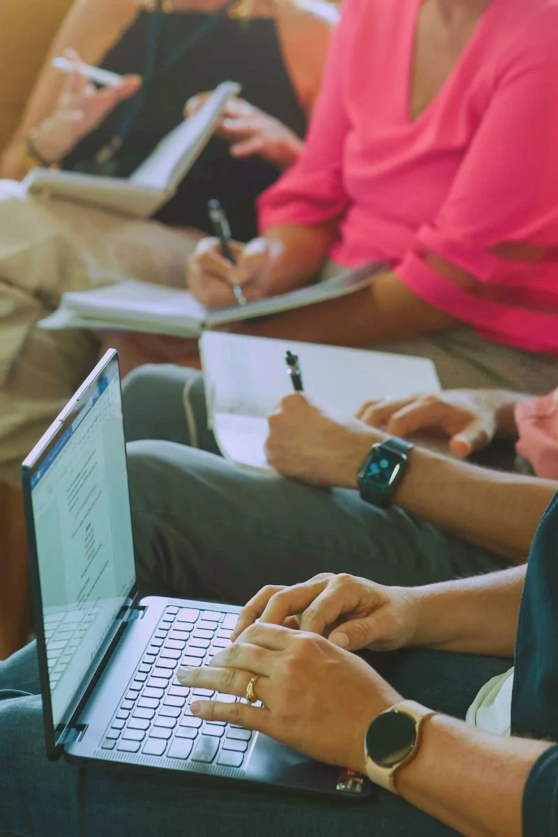People sitting and taking notes during a meeting or workshop, with some using laptops and others writing in notebooks.