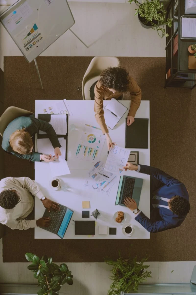 Top-down view of five people working at a white table with laptops, documents, charts, and coffee cups in a modern office.