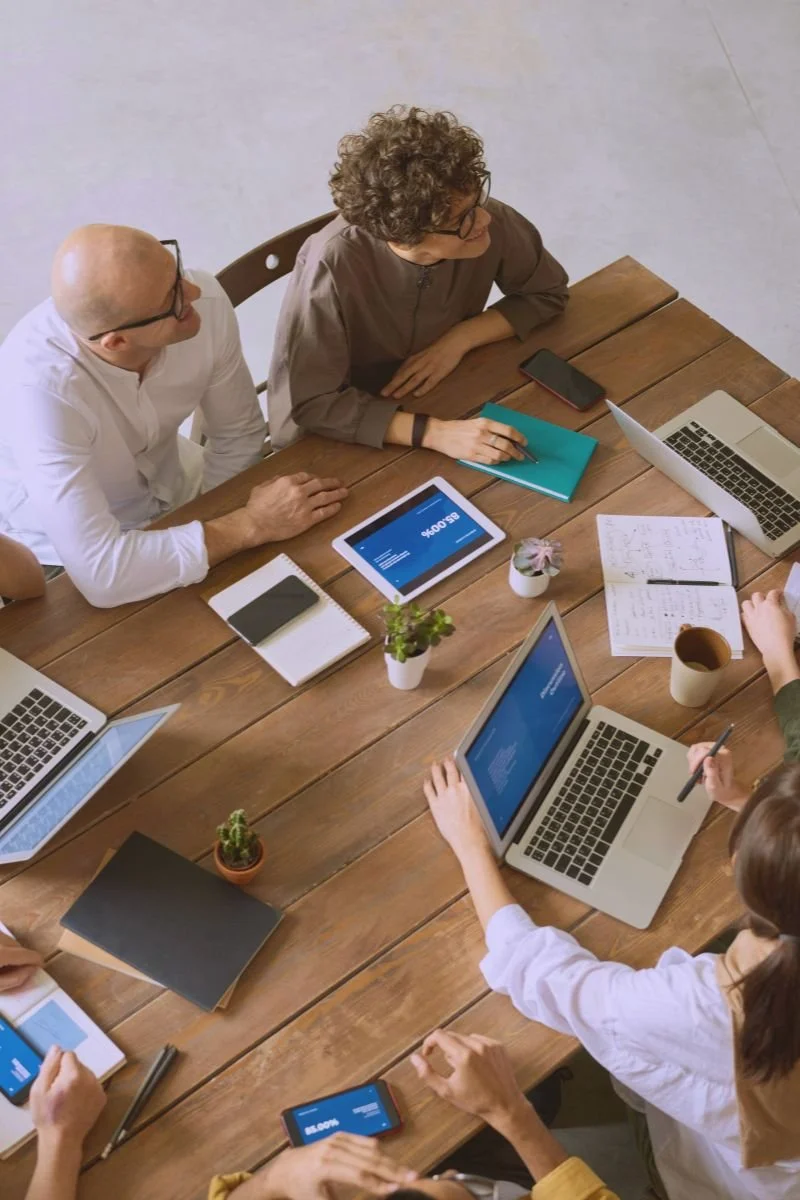 A group of people gathered around a wooden table during a meeting, working on laptops, tablets, and smartphones.