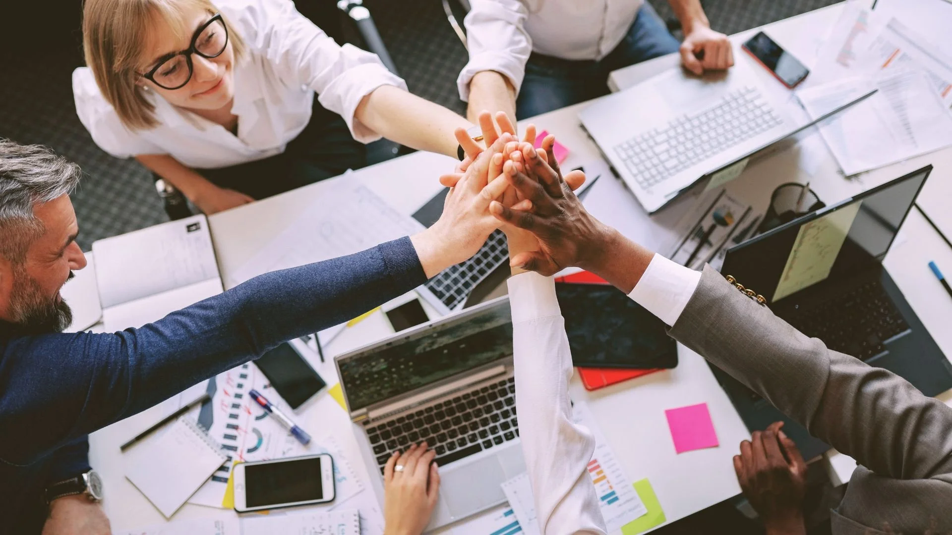 A diverse group of five people in a business meeting, giving each other a group handshake over a cluttered table with laptops, documents, and electronic devices.