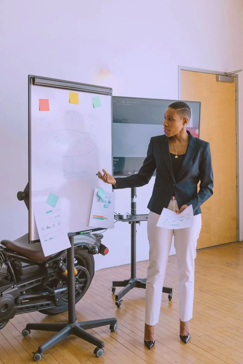 A woman in a black blazer and white pants giving a presentation in a room with a brown door, a whiteboard with sticky notes, and a motorcycle placed next to the whiteboard.