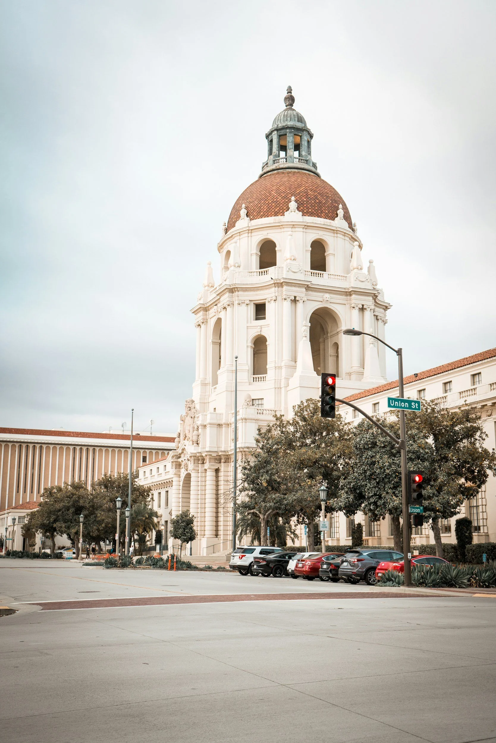 A historic white building with a large red-domed tower and a smaller cupola on top, surrounded by traffic lights, street signs, parked cars, trees, and a wide street, under an overcast sky.