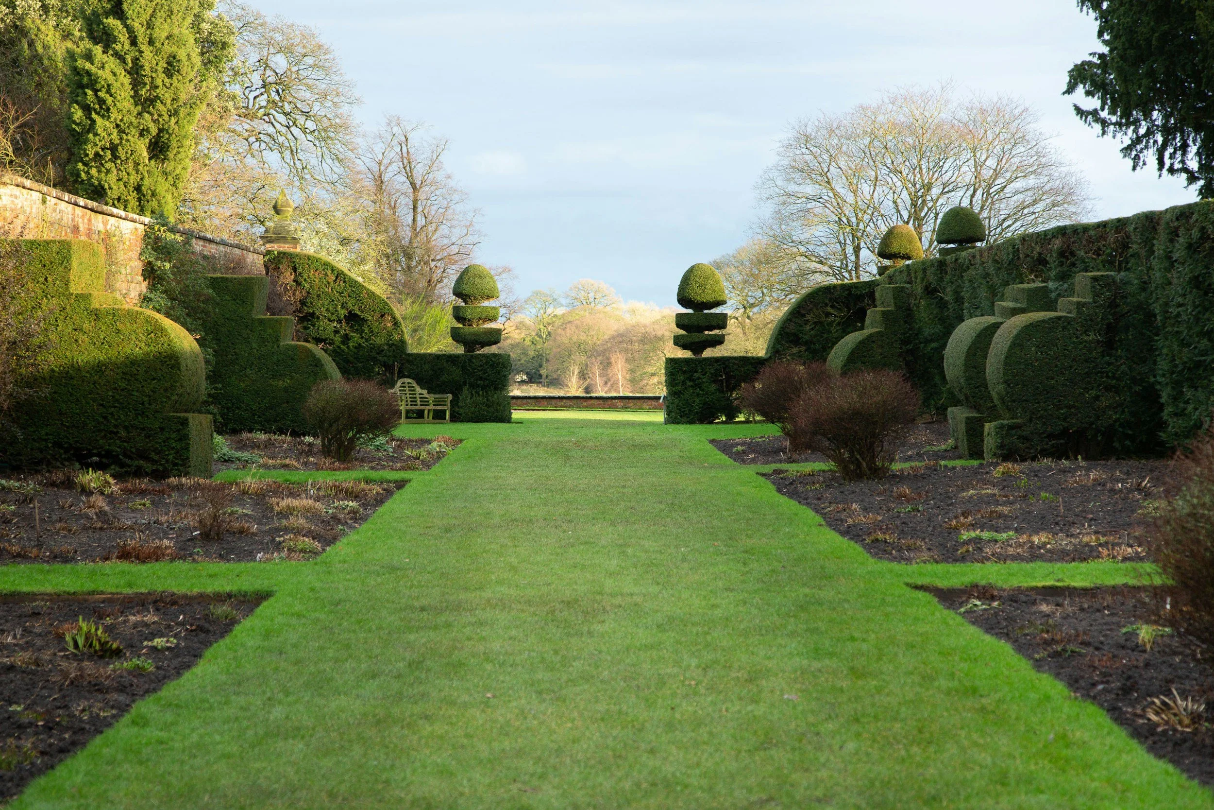 A manicured garden with a green lawn, surrounded by trimmed hedges and topiary trees, with a backdrop of leafless and evergreen trees and a stone wall.