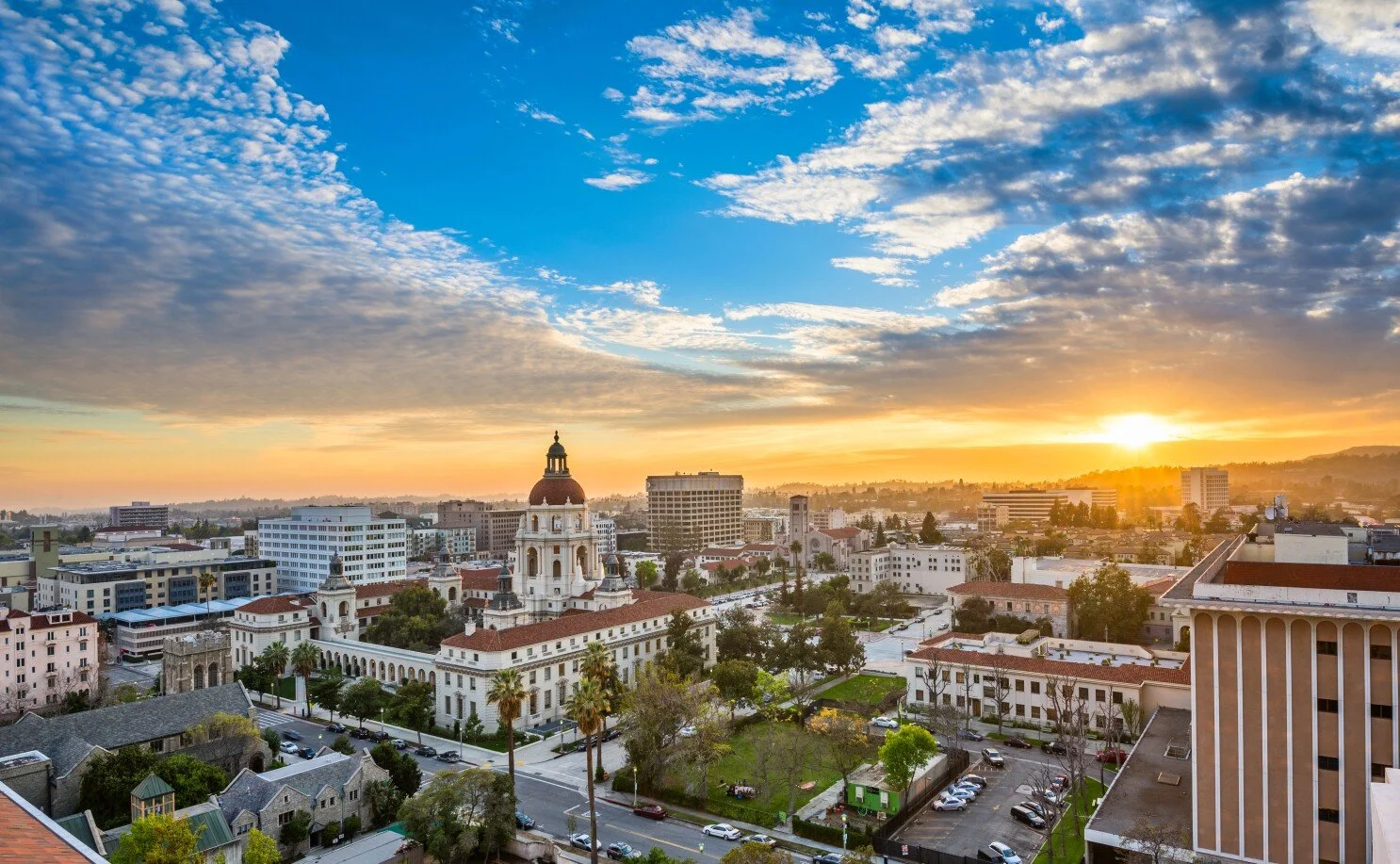 Sunset over a cityscape featuring historic and modern buildings, a prominent church with a dome, and a green park in the foreground.