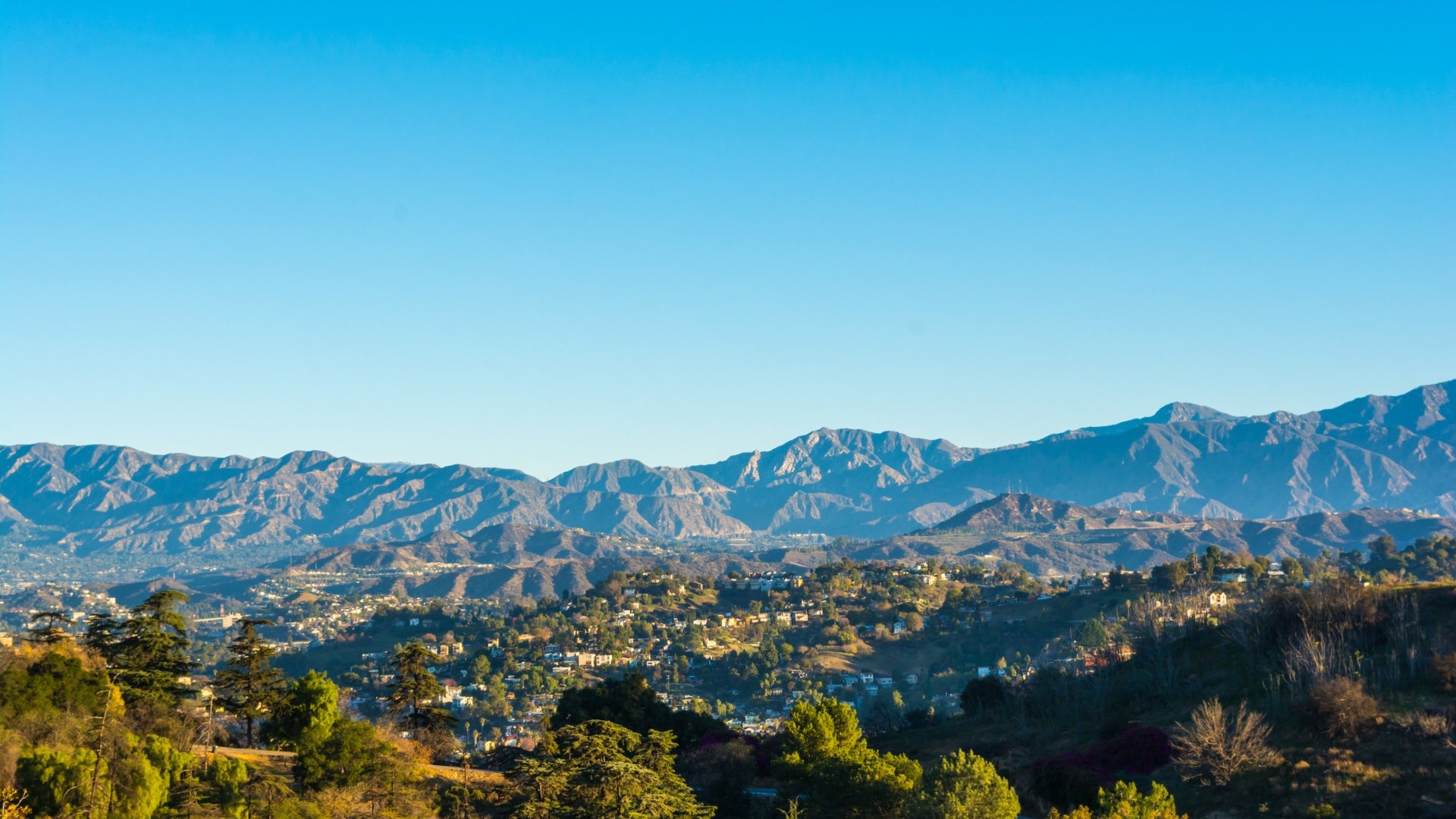 A mountain range with clear blue sky above and a city in the foreground surrounded by trees.