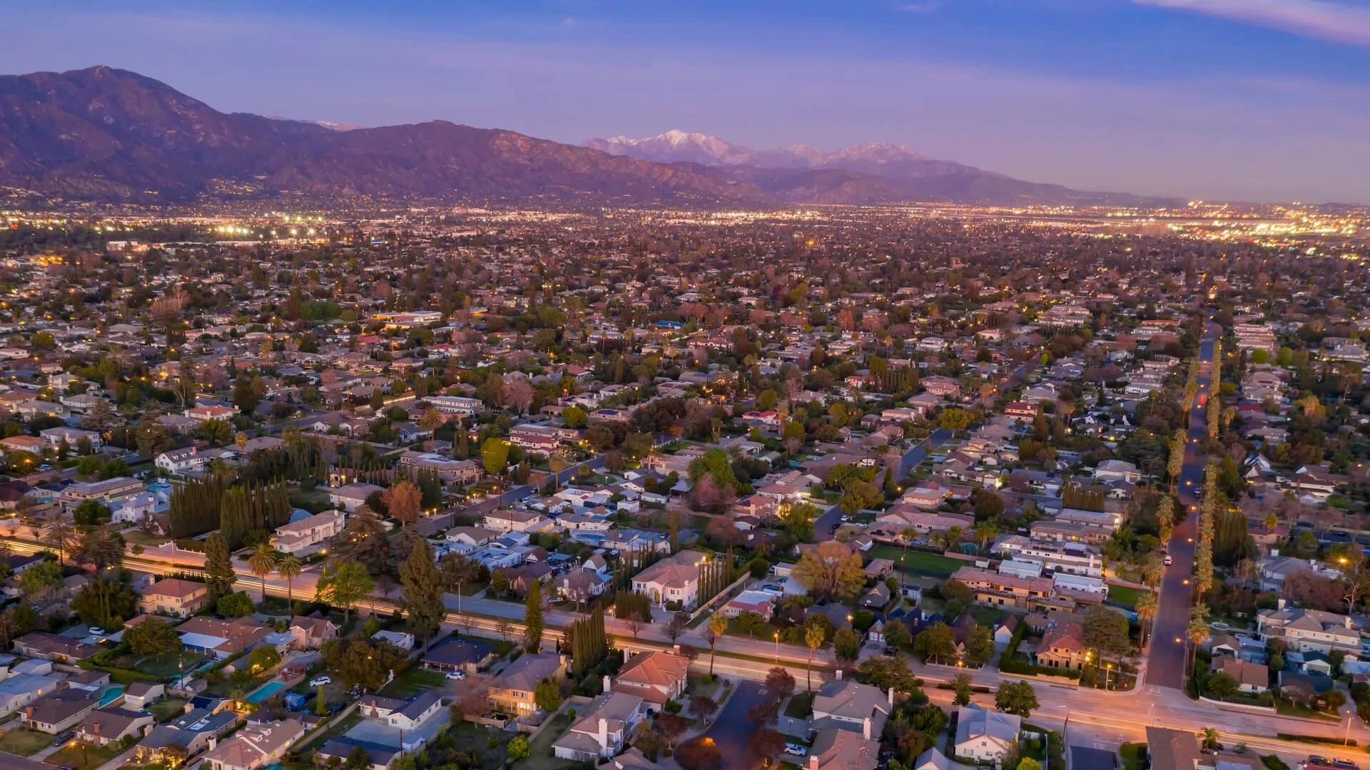Aerial view of a city at dusk with mountain range in the background and houses lit by streetlights in the foreground.