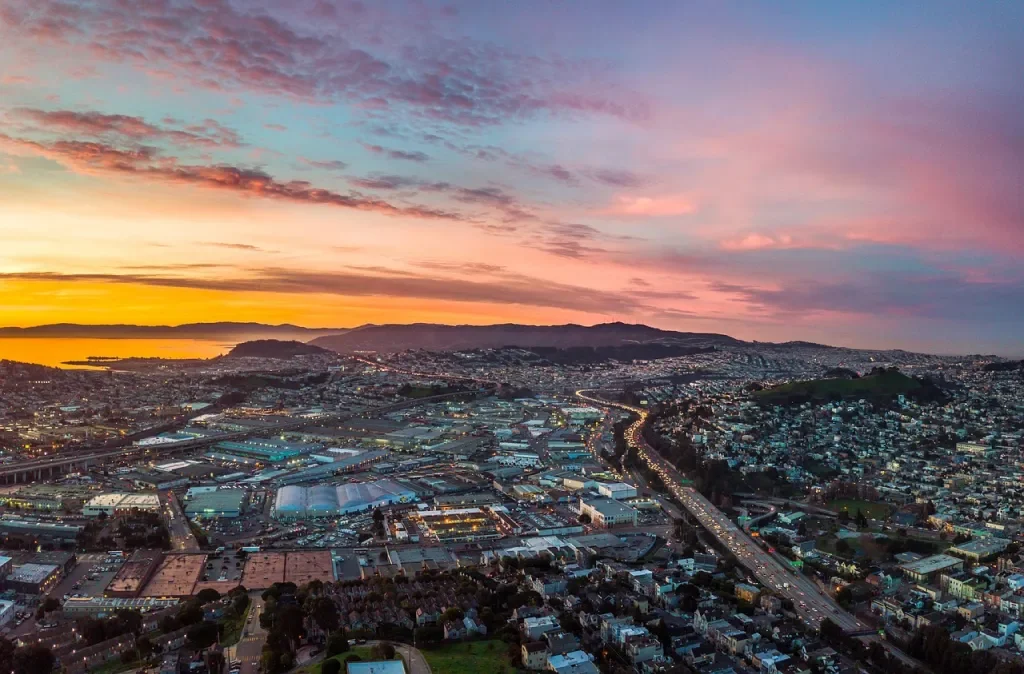 Aerial view of a city at sunset with colorful sky, buildings, roads, and hills in the background.