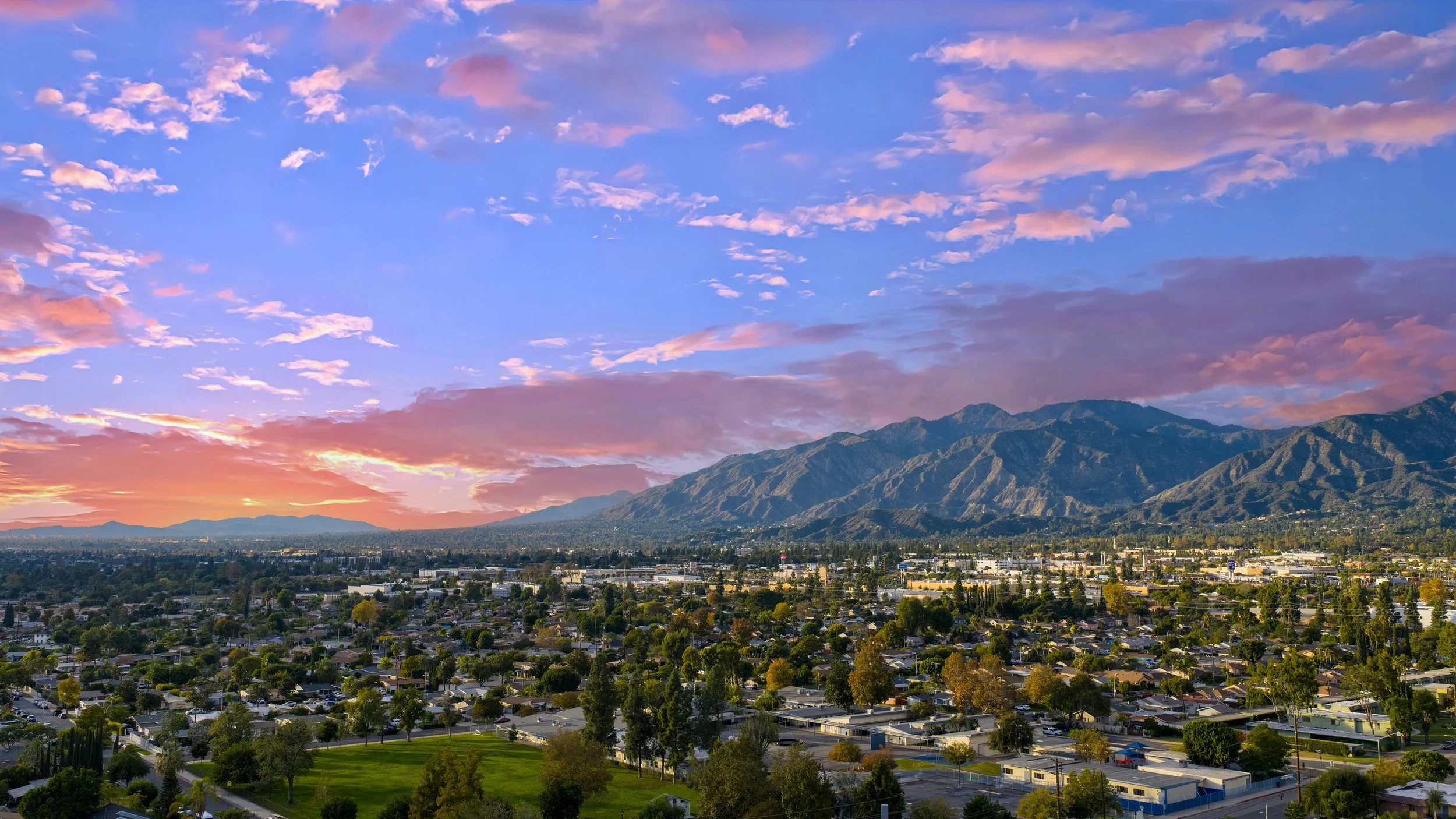 View of a cityscape with a mountain range in the background during sunset. The sky is filled with pink and purple clouds.
