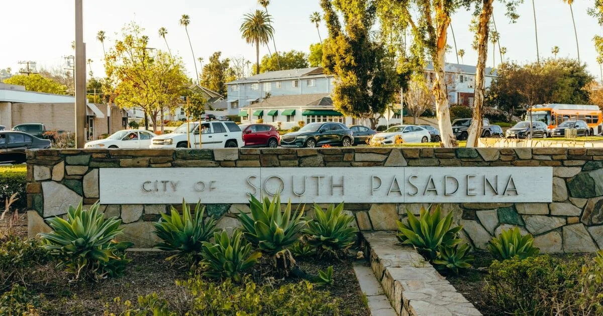 A stone sign with the text 'City of South Pasadena' in front of a parking lot with cars and buildings, surrounded by trees and plants.