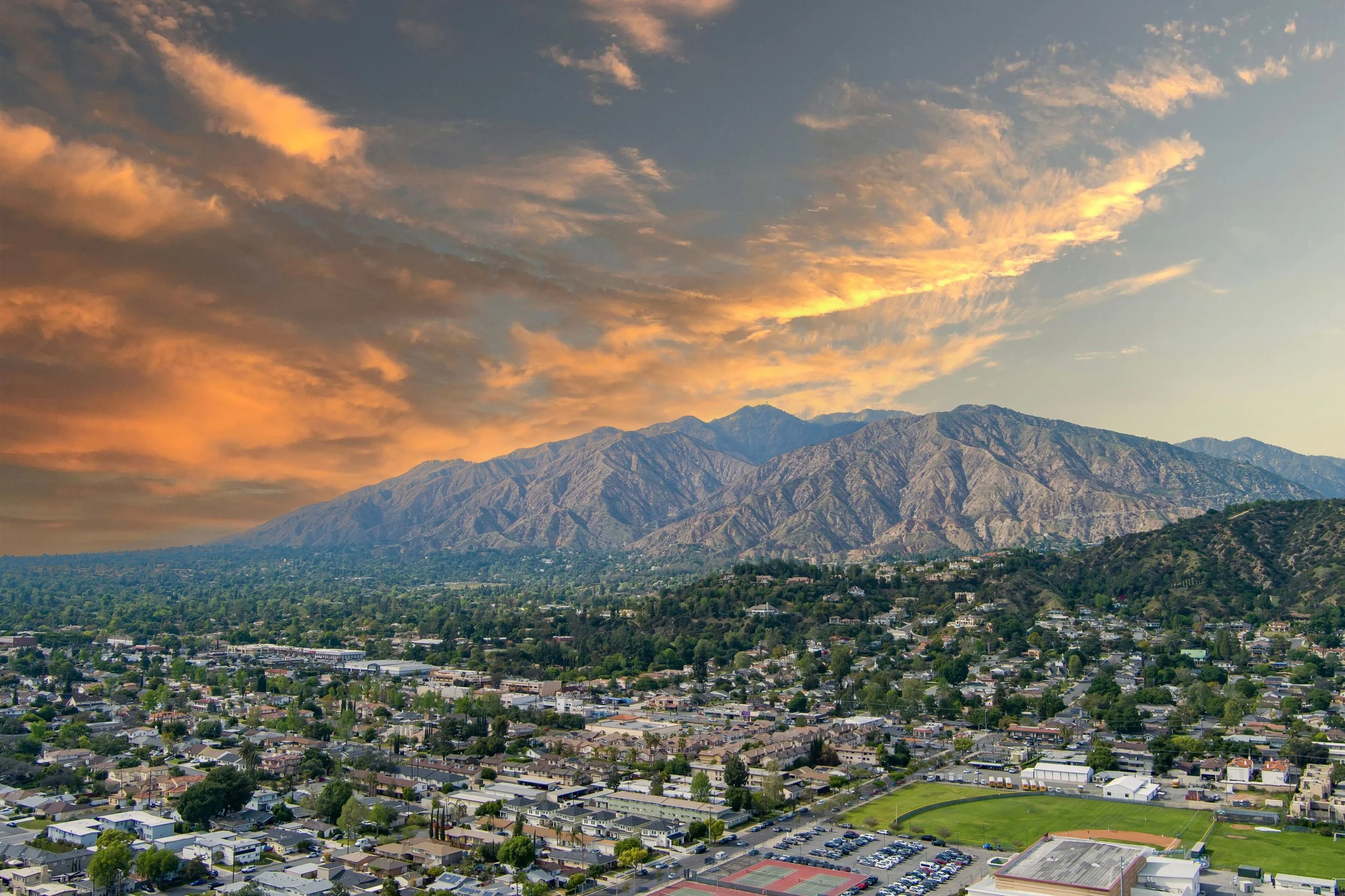 A cityscape with residential houses, a baseball field, and a parking lot in the foreground. Mountains under a partly cloudy sky with orange and pink clouds at sunset in the background.