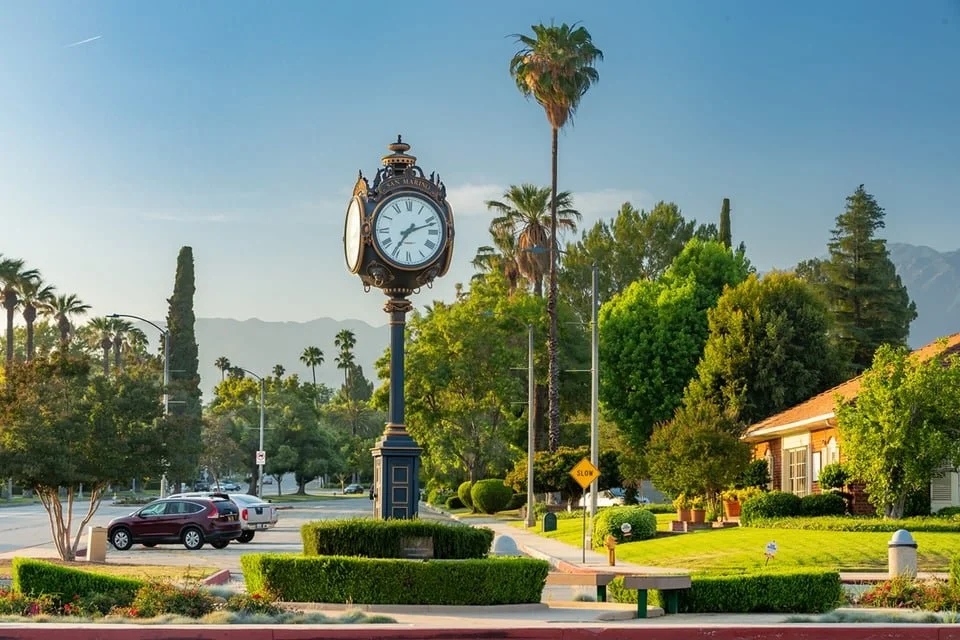 A vintage-style clock tower on a sidewalk in a small town, surrounded by trees, houses, and parked cars, with mountains in the background on a clear day.