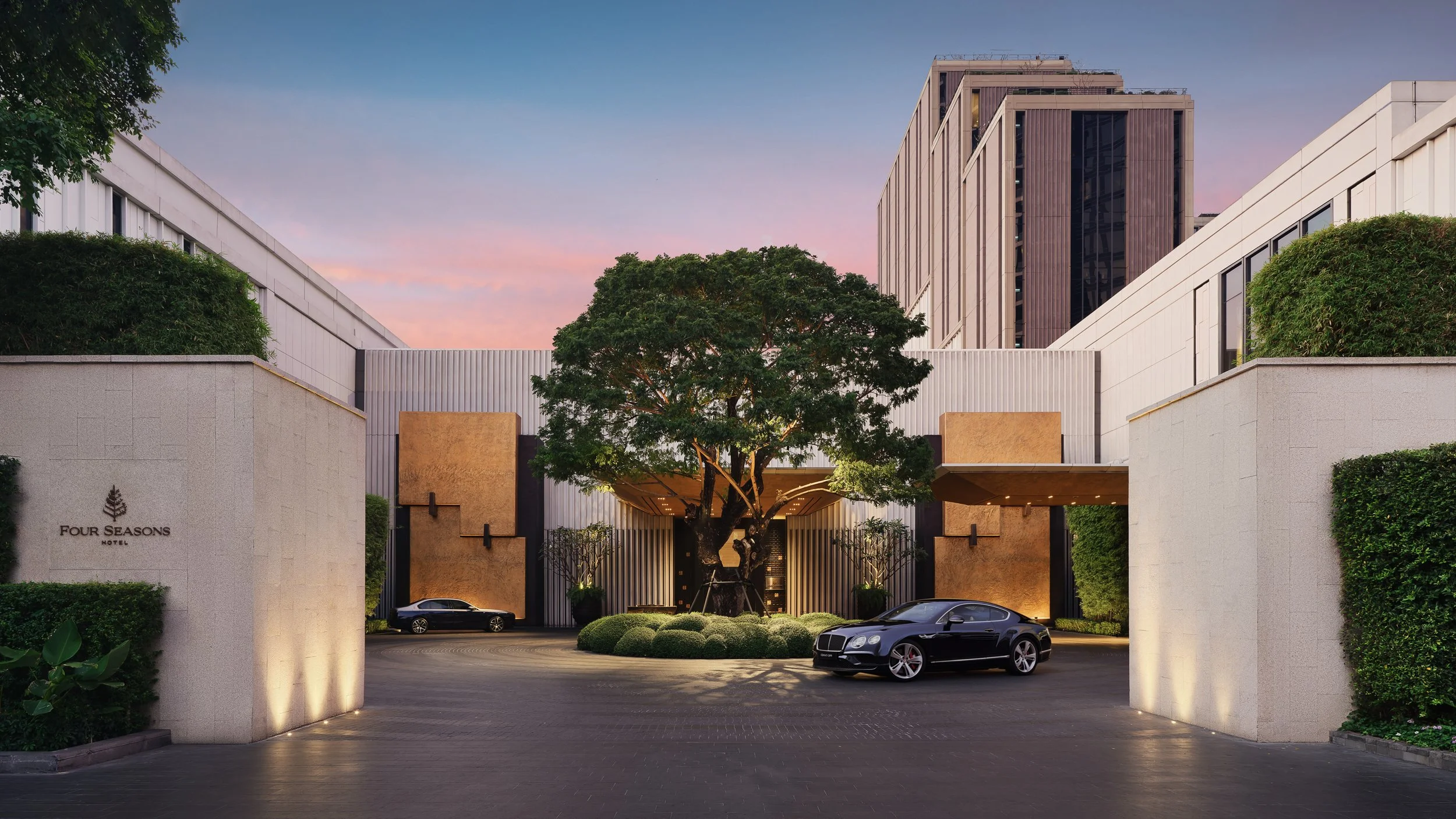 Luxury hotel entrance with modern architecture, large tree in the center, and luxury cars parked outside at dusk in Bangkok.