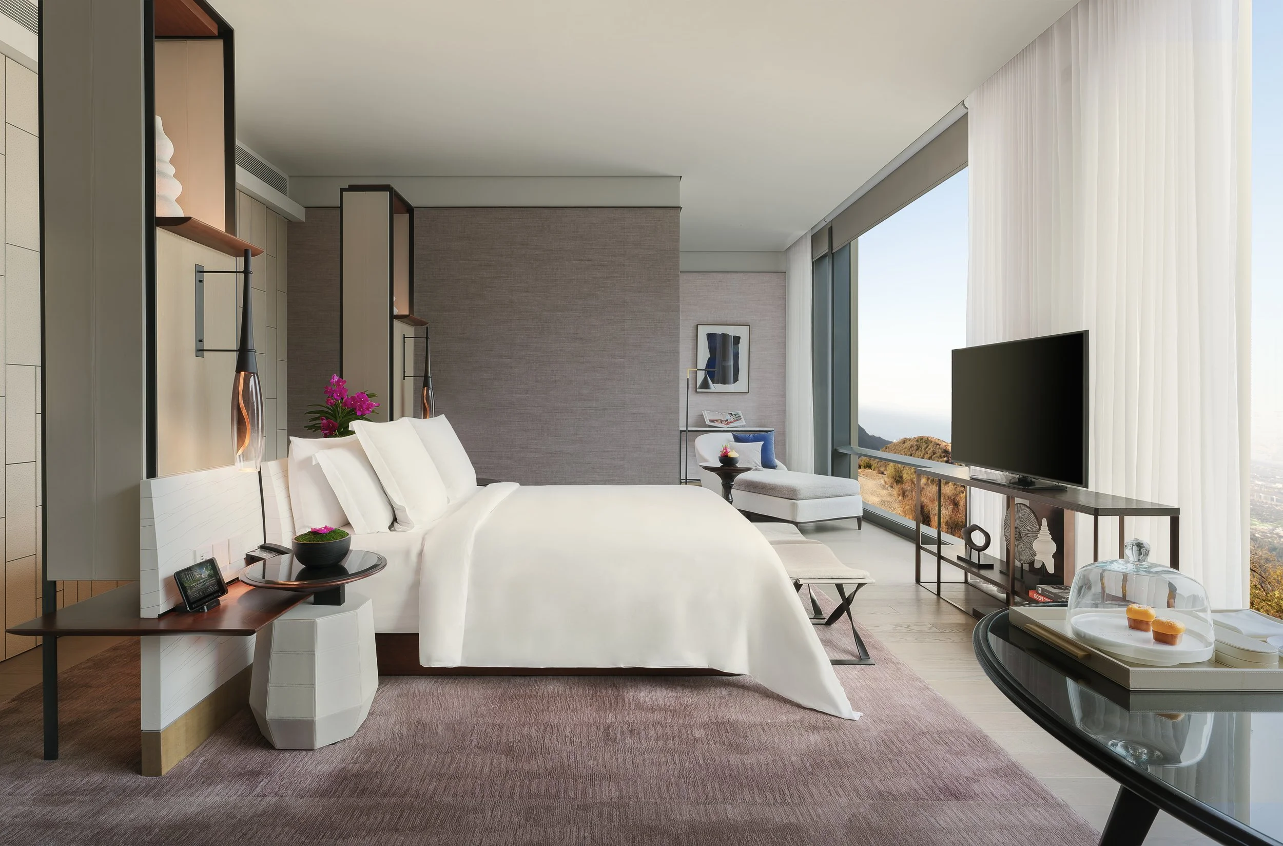 Luxury hotel suite featuring a large bed with white bedding, upscale decor, a flat-screen TV, a lounge chair with a blue cushion, and a panoramic window with a scenic view of Bangkok's main river.