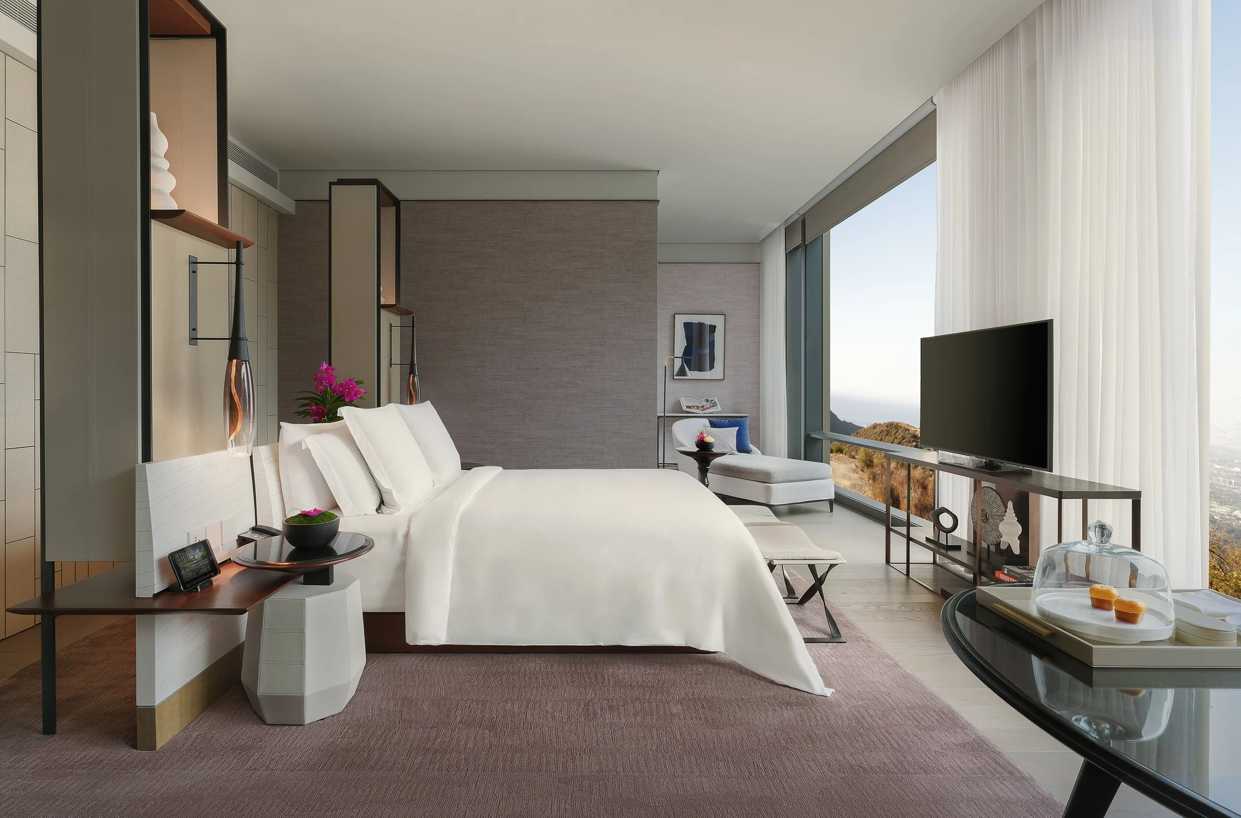 Luxury hotel suite featuring a large bed with white bedding, upscale decor, a flat-screen TV, a lounge chair with a blue cushion, and a panoramic window with a scenic view of Bangkok's main river.