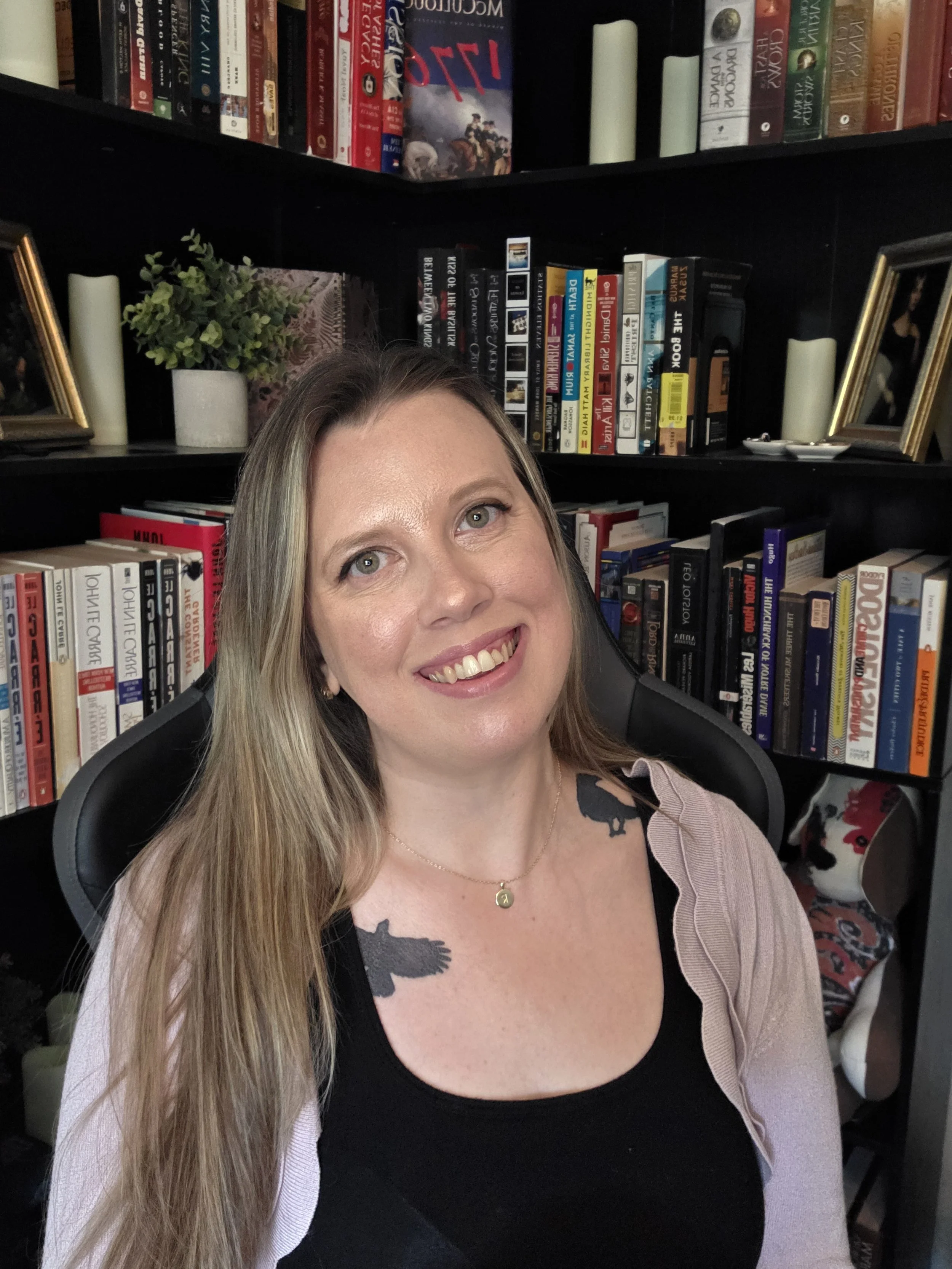 A smiling woman with long blonde hair sitting in a black chair, with a bookshelf behind her filled with books, framed photographs, candles, and decorative items.