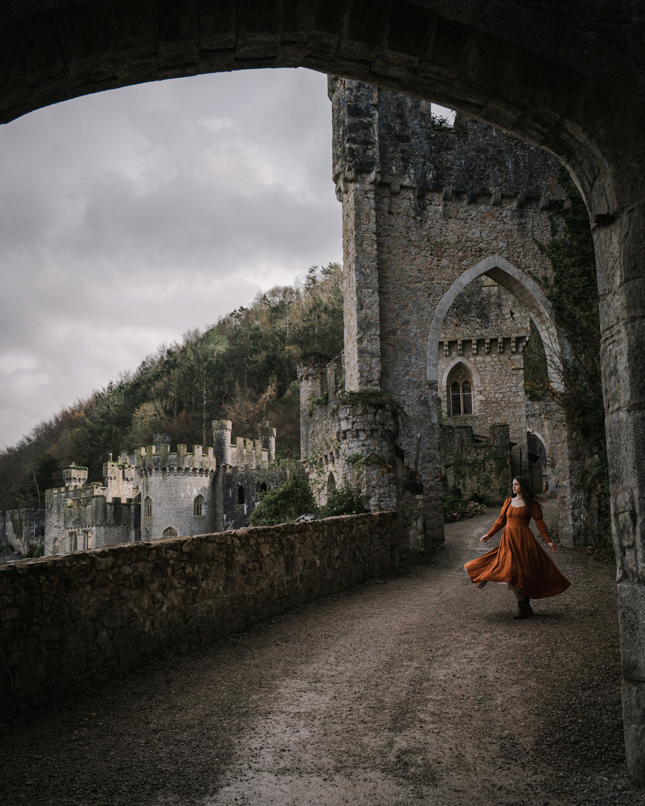 A woman in a long, flowing, rust-colored dress twirling outdoors beneath an ancient stone castle archway with a cloudy sky overhead and lush greenery on a hillside in the background.