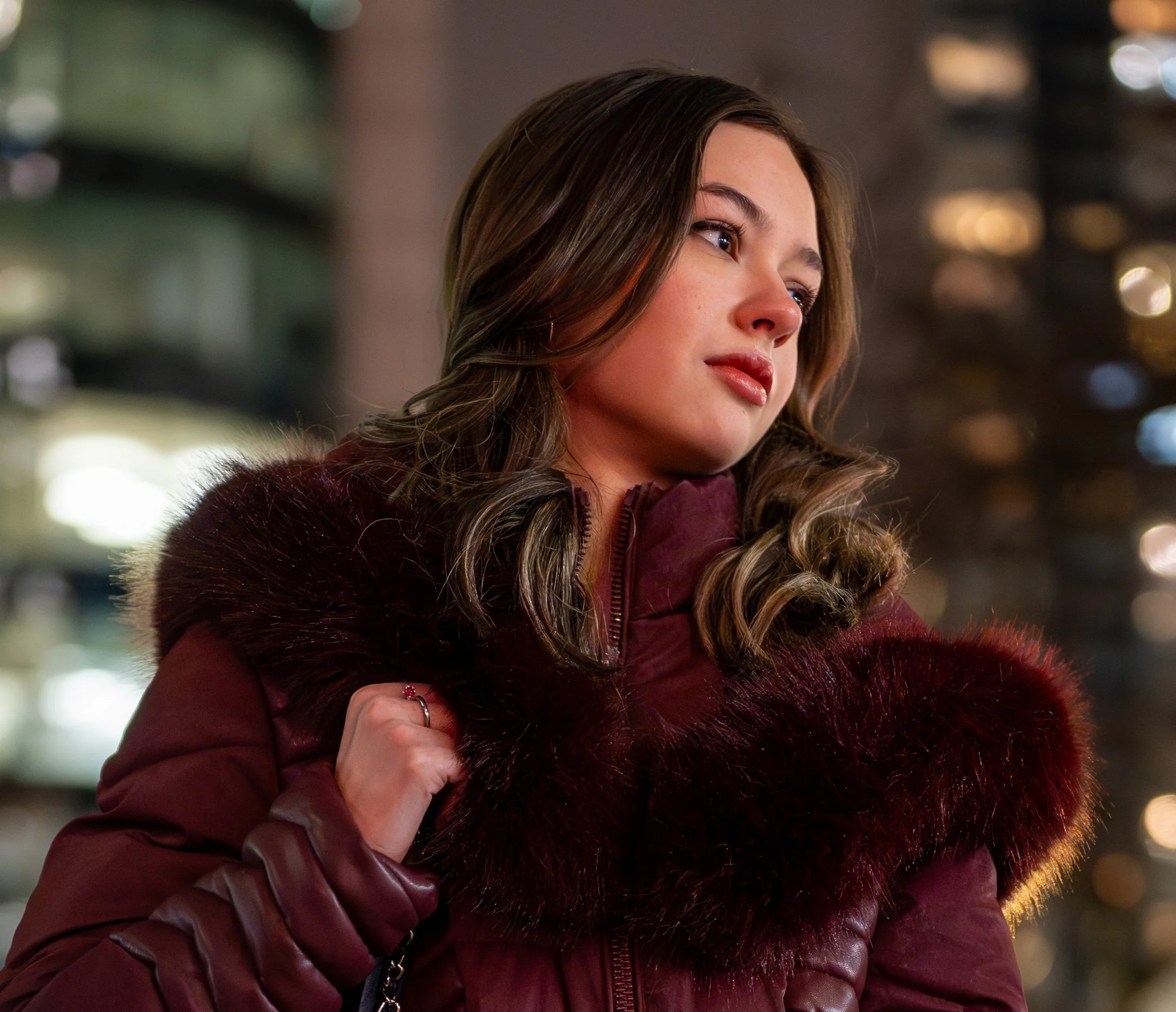 A young woman with brown hair wearing a maroon winter coat with fur trim, standing outdoors at night with blurred city lights in the background.