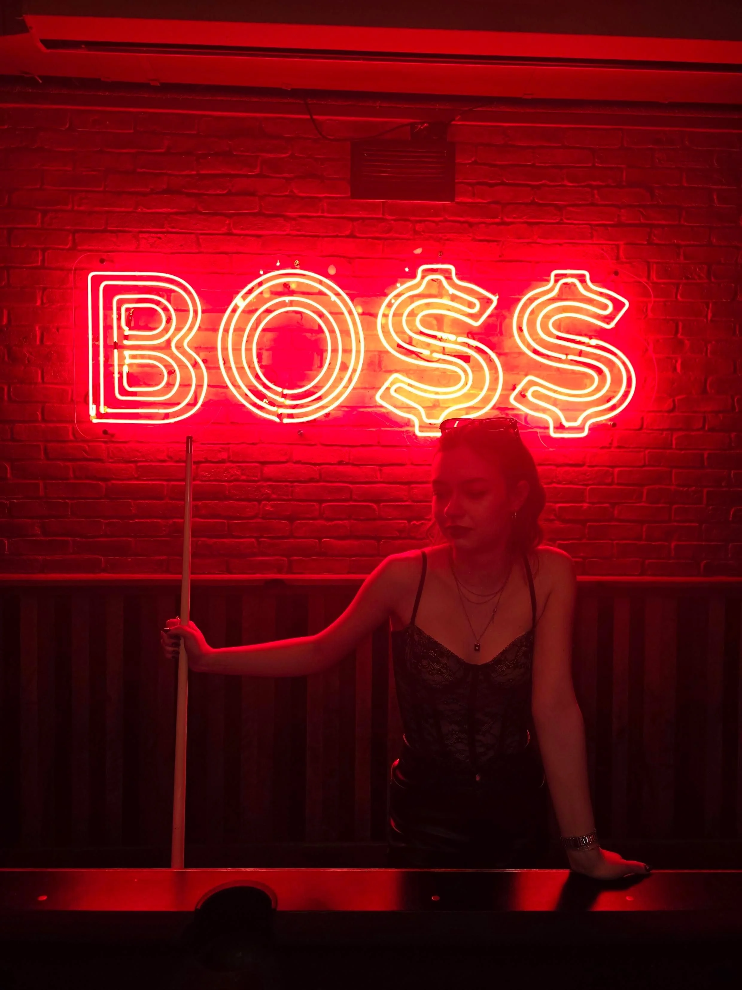 A woman in a black lace top posing in front of a red neon sign that says 'BOSS' with dollar signs replacing the 'S's'